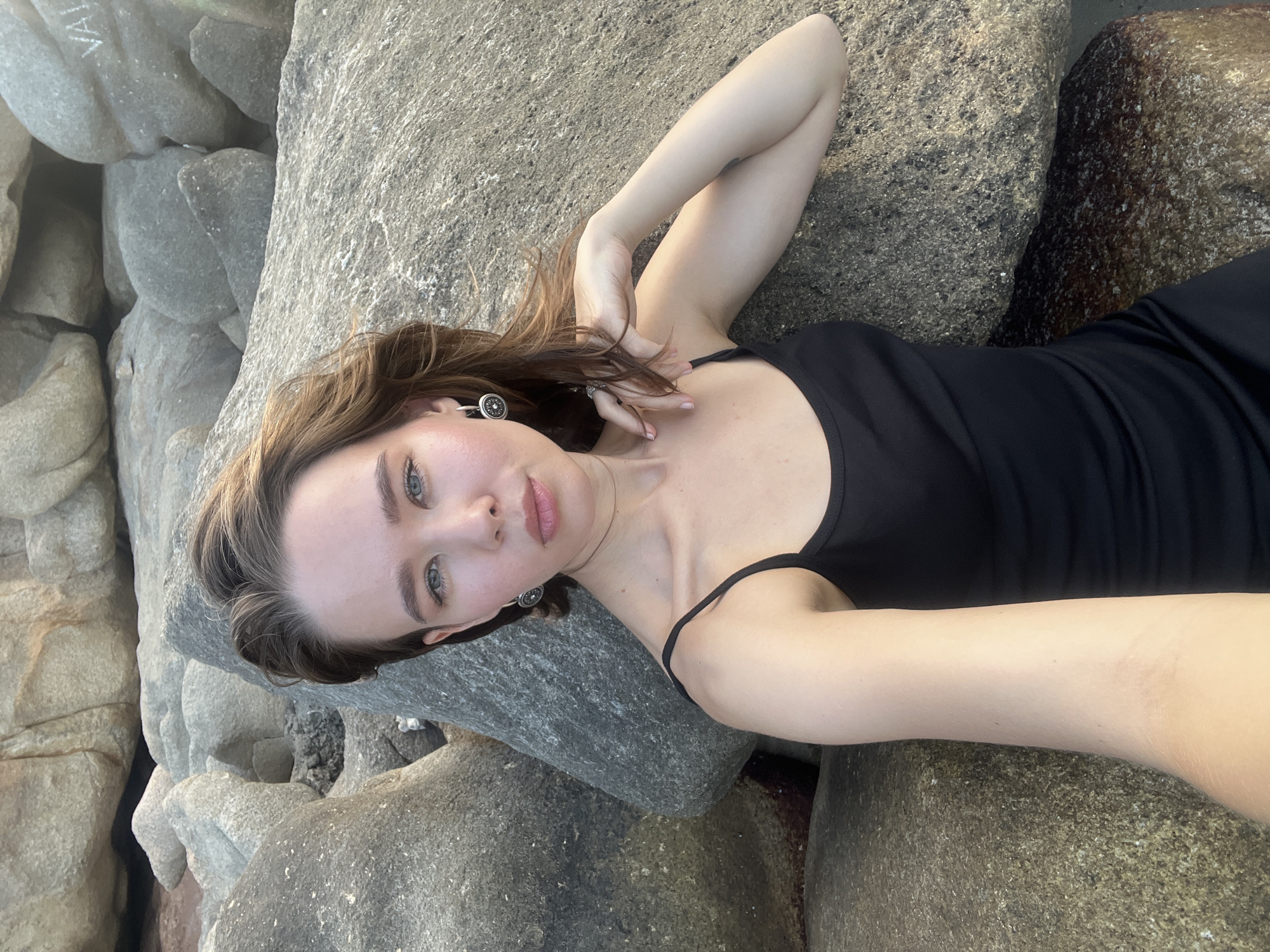 A young woman with light brown hair and a calm expression is depicted lying on large, grey rocks. She is wearing a black spaghetti strap top or dress and looks directly at the camera. Her right arm is bent with her hand gently touching her hair, and a distinctive circular earring is visible on her left ear.
The setting is an outdoor rocky environment, consistent with a coastal or beach area like Bac Nha Trang Ward, Vietnam. The rocks vary in size and texture, creating a natural backdrop. The lighting is soft and natural, suggesting daytime, possibly late morning or afternoon, with no harsh shadows. On one of the rocks in the upper left portion of the image, the word "VALE" is faintly carved or written. The woman's pose is relaxed, and her expression is serene. The composition suggests a close-up shot, likely a selfie, with the woman positioned diagonally across the frame.