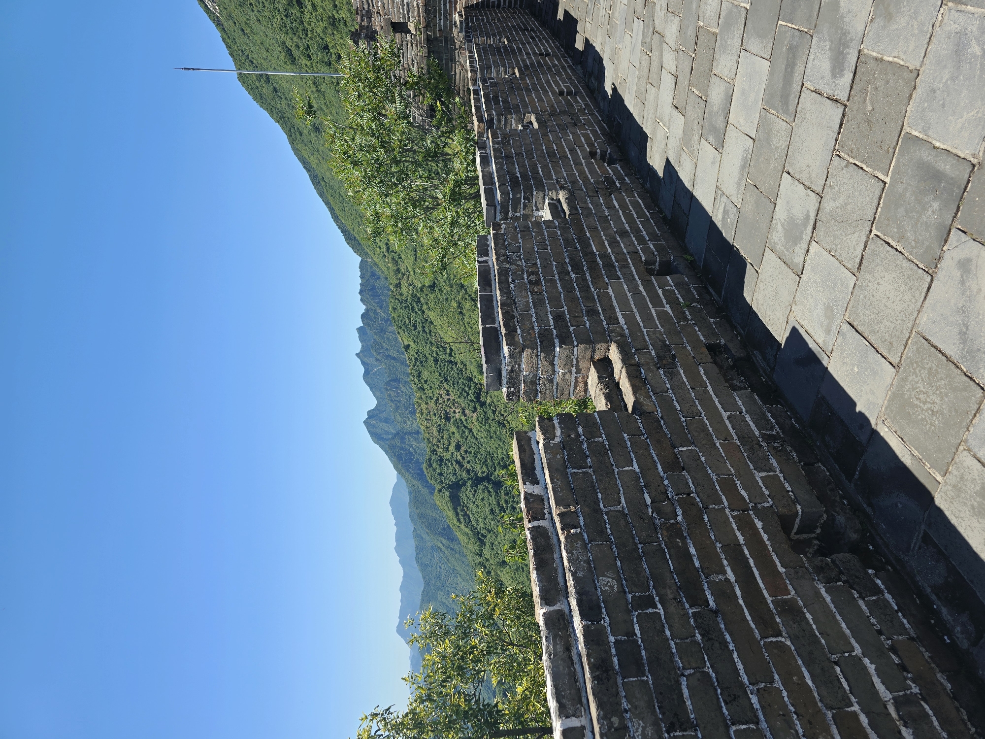 The image depicts a section of the Great Wall of China, likely located in Huairou District, China, given the mountainous, forested terrain. In the foreground, the weathered grey brickwork of the wall is prominent, with a paved walkway of large, grey rectangular stones on its right. The wall features crenellations and what appear to be embrasures. Beyond the wall, rolling green mountains covered in dense vegetation dominate the middle ground, stretching into the distance under a clear, vibrant blue sky. A tall, slender flagpole is visible on the crest of the mountain to the right, topped with a pointed object. The lighting suggests it is a bright, sunny day, casting distinct shadows on the walkway. No people are visible in the frame.