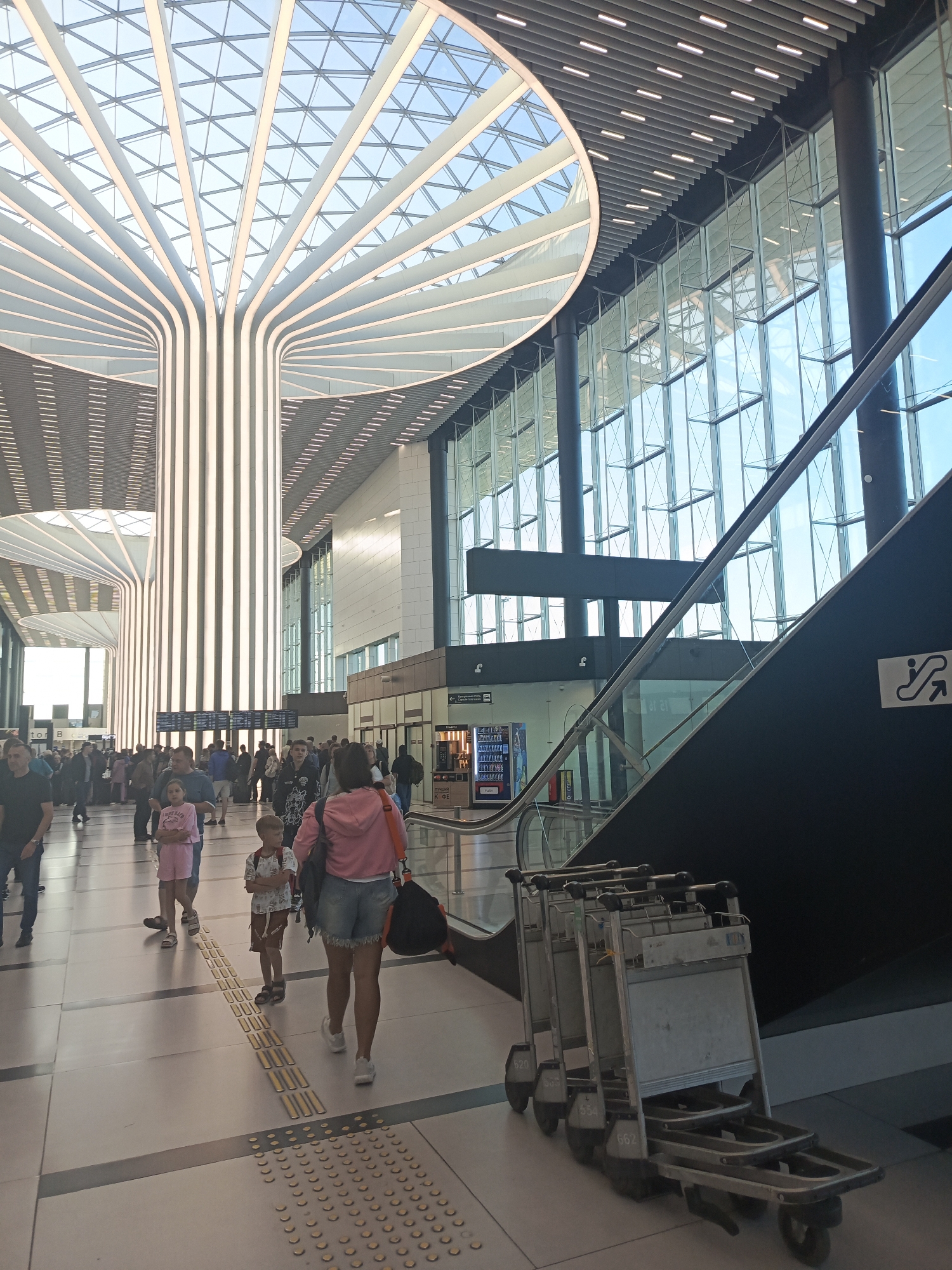 The image depicts the interior of a modern airport terminal, likely at the airport in Ob, Russia, given the context provided. The terminal features a striking architectural design with a high, vaulted ceiling composed of glass and metal, allowing natural light to flood the space. Several large, vertically ribbed structures, illuminated from within, extend from the floor towards the ceiling, creating a visually dramatic effect.
In the foreground, a group of people are walking through the terminal. A woman in a pink hooded jacket and denim shorts is walking away from the camera, carrying a black bag. She is accompanied by a young girl in pink shorts and a t-shirt. To their left, a young boy in a patterned shirt and shorts stands with his arms crossed. Further in the background, a crowd of travelers are visible, some with luggage, suggesting ongoing travel activities.
To the right of the frame, a dark escalator ascends, its metal railing and glass shielding clearly visible. Beside the escalator, a sign with an escalator icon is mounted on the wall. In the middle ground, a row of luggage carts are neatly stacked and ready for use. Vending machines are also present in the background, along with departure boards displaying flight information. The floor is tiled and features tactile paving for visually impaired individuals. The overall impression is one of a busy, contemporary transportation hub.