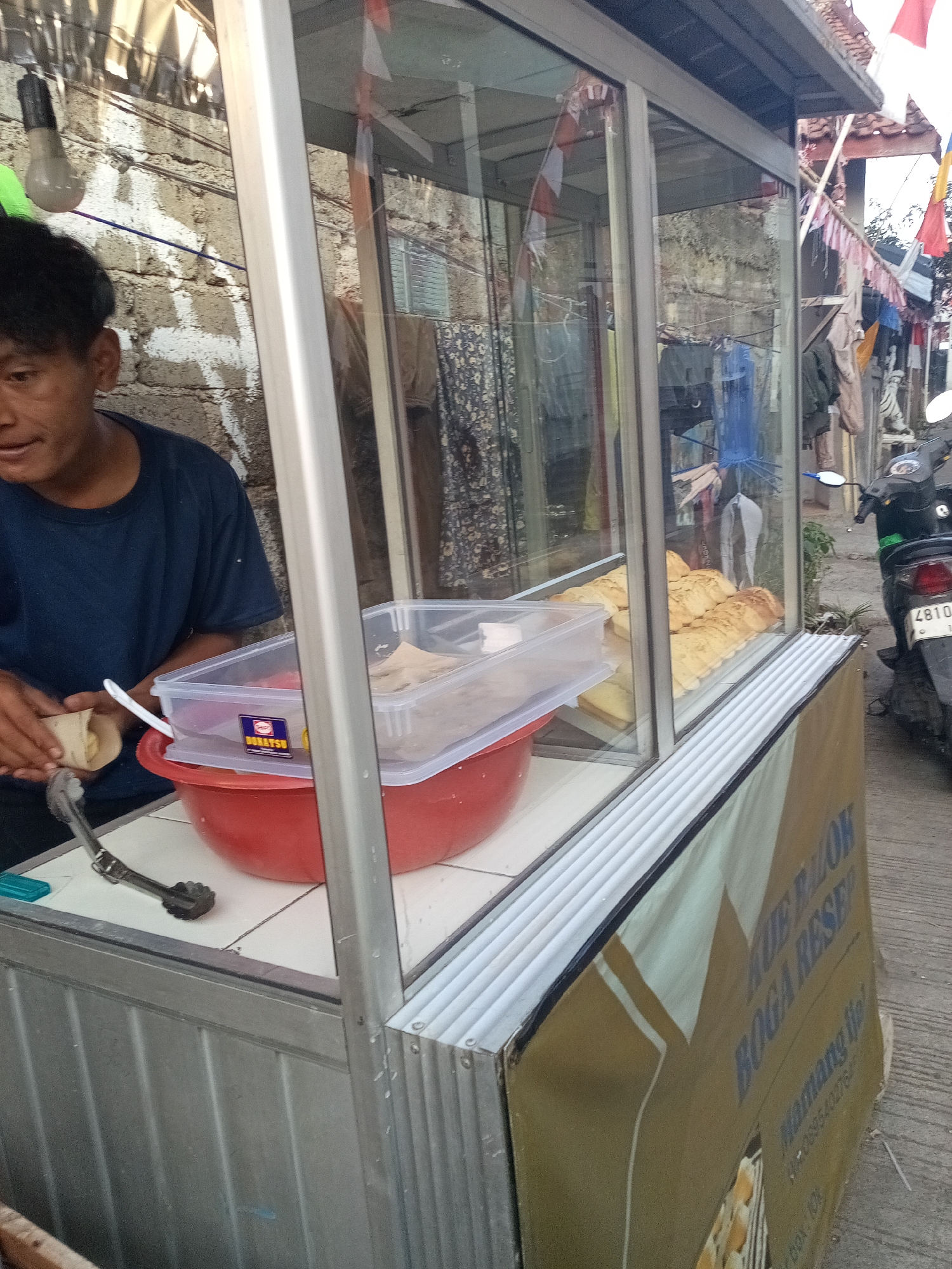 In Cihanjuang, Indonesia, a vendor, a male with dark hair and a navy blue t-shirt, is preparing food at a food stall. He is holding a rolled pastry in his left hand, with his right hand partially obscuring it. The stall features a glass display case showcasing several golden-brown pastries. To the left of the stall, a red bowl sits on a tiled surface, with a clear plastic container filled with more food items placed above it. Tongs rest near the bowl. The background shows a brick wall and various items hanging from a clothesline, suggesting an outdoor market or street vendor setup. A scooter is parked to the right of the stall. The stall has a banner with text that appears to be in Indonesian.