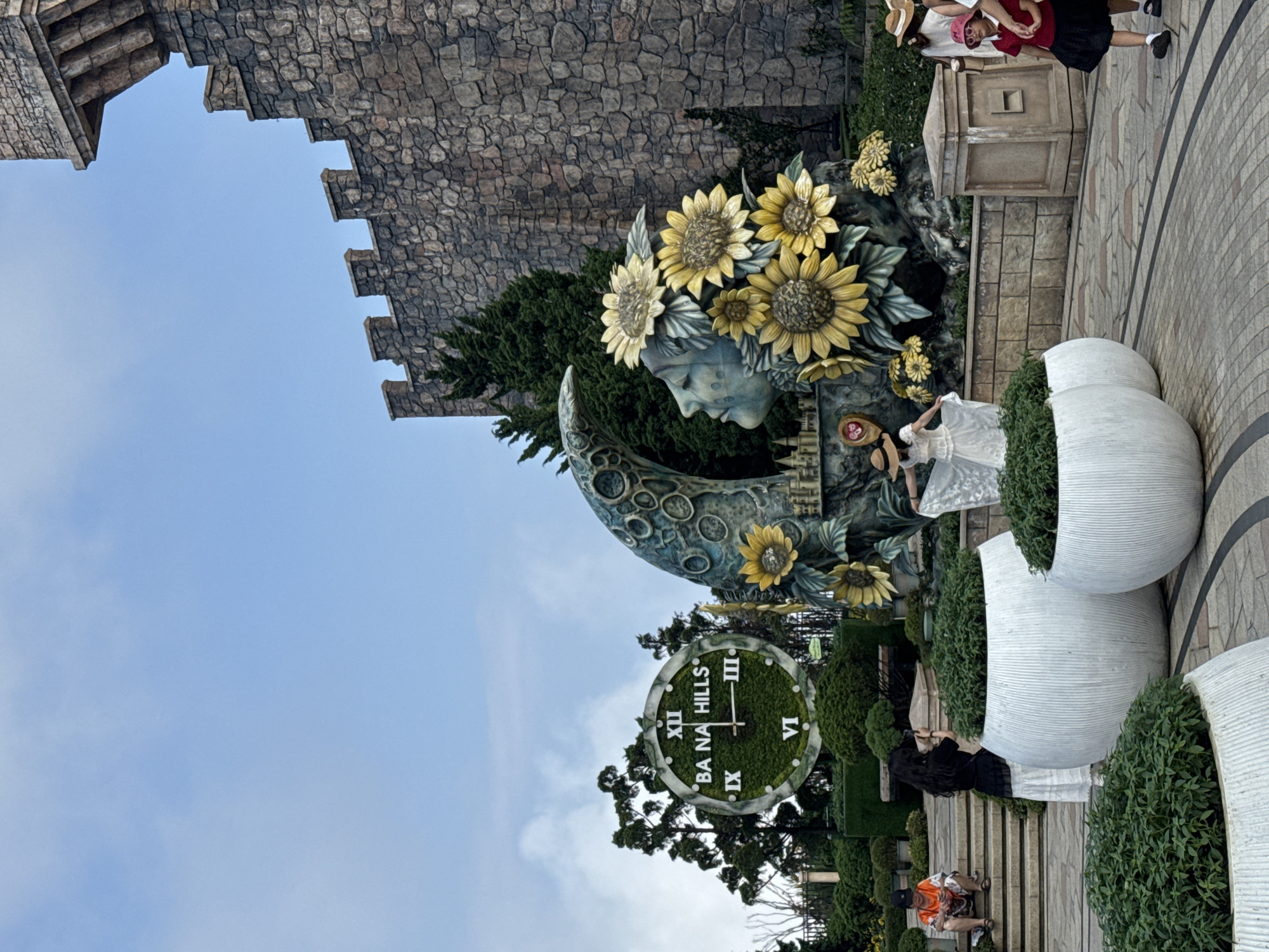 The image captures a scene in what appears to be Ba Na Hills, a mountain resort near Đà Nẵng, Vietnam. The sky is partially visible with a mix of blue and overcast. A medieval-style stone wall looms in the background, possibly part of a faux-historic structure within the resort.
The focal point is a sculpture of a face crowned with sunflowers, emerging from a crescent moon. Below this, there's a large green clock with white Roman numerals and the words "Ba Na Hills" inscribed on it.
Several people are present. A woman in a white dress poses near the sculpture. Another group of people can be seen further down, with one person sitting on the stairs. Two other women in a dress and a woman taking a picture.
The environment includes large, decorative white planters filled with green bushes, contributing to a stylized, garden-like atmosphere. The ground is paved with gray paving stones. The scene appears to be a tourist spot with carefully curated landscaping and thematic architecture.