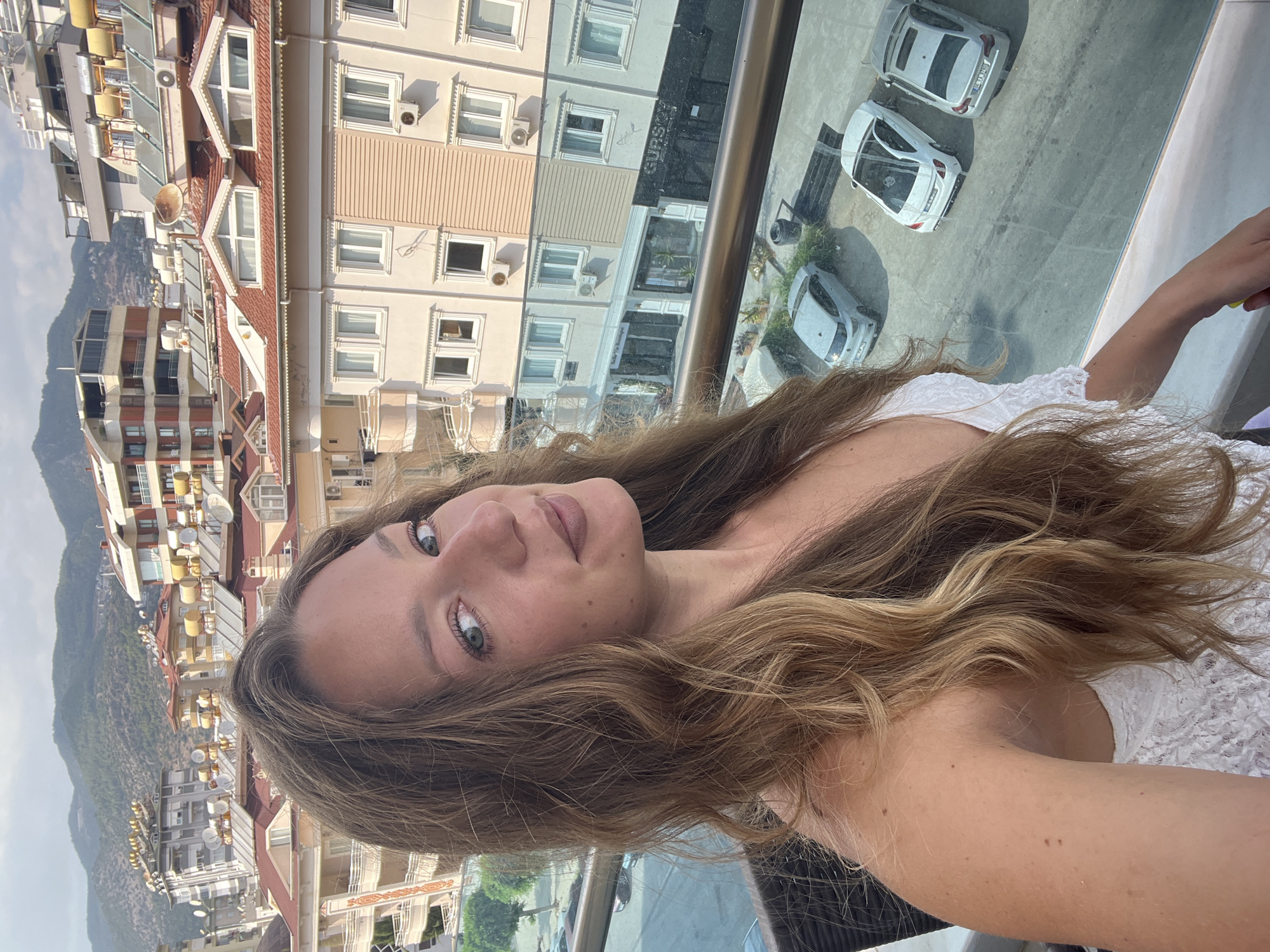 In Alanya, Turkey, a woman with long, wavy brown hair and light green eyes, wearing a white lace top, poses for a selfie from a balcony overlooking a street with parked cars and buildings. The buildings feature red-tiled roofs, solar water heaters, and air conditioning units. The woman's facial features are clear, including her nose, lips, and a scattering of freckles across her cheeks. The background shows a mountainous landscape and the urban sprawl of Alanya.