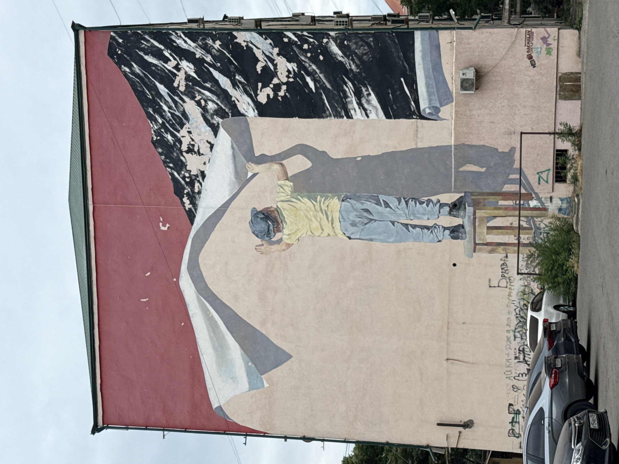 This is a photograph of a mural painted on the side of a building. The mural depicts a man in a yellow shirt and blue pants, wearing a blue cap. He is lying down and appears to be unrolling a large sheet of paper or canvas, which is colored a light beige. The top portion of the mural features abstract black and white shapes, possibly representing a torn or peeling surface. The man is casting a shadow on the beige surface.
The scene is set outdoors, with a cloudy sky visible above the building. There is an air conditioning unit mounted on the wall of the building, to the right of the man's head. Below the mural, several cars are parked along a street. The cars are dark-colored, including a white SUV and several darker sedans. Green bushes and trees are visible on the right side of the frame, growing near the building and the street.
Graffiti and text are visible on the wall of the building, both within and outside the painted mural. Some of the text appears to be in Cyrillic script, including words like "БРАВА" and "ОЧАЛАВ GROUP". There are also some illegible markings and tags. The overall impression is that of an urban setting with public art. The lighting suggests it might be daytime, possibly overcast. The mural itself conveys a sense of work or creation, with the man engaged in the act of revealing or preparing a large surface.