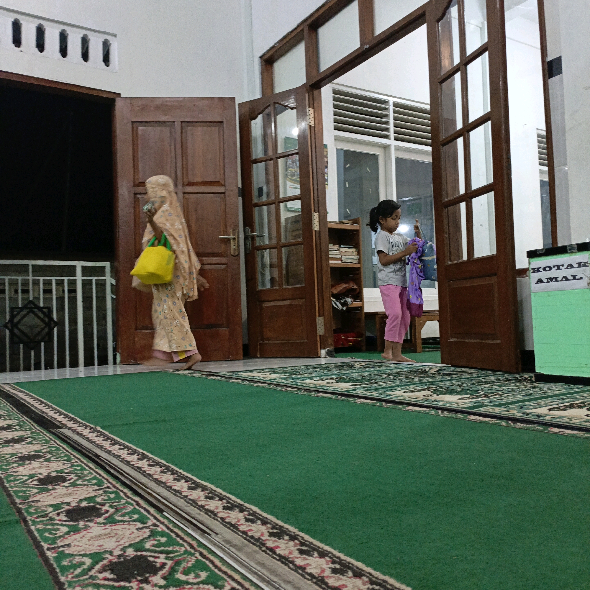 An outdoor view of the entrance to a building in Salatiga, Indonesia, where two individuals, likely of Indonesian ethnicity, are present. The first individual, a woman, is walking away from the camera and towards a closed wooden door on the left. She is wearing a patterned headscarf and garment, and carries a bright yellow bag with green straps over her right shoulder. The second individual, a young girl, is standing inside the building, facing the camera and holding a purple garment. She is wearing a grey t-shirt and pink pants and appears to be barefoot. The floor is covered with green carpeting featuring a patterned border. The building's exterior shows white walls and wooden doors and window frames. To the right of the girl, a green donation box with the text "KOTAK AMAL" is visible. The lighting suggests it is evening or dusk.