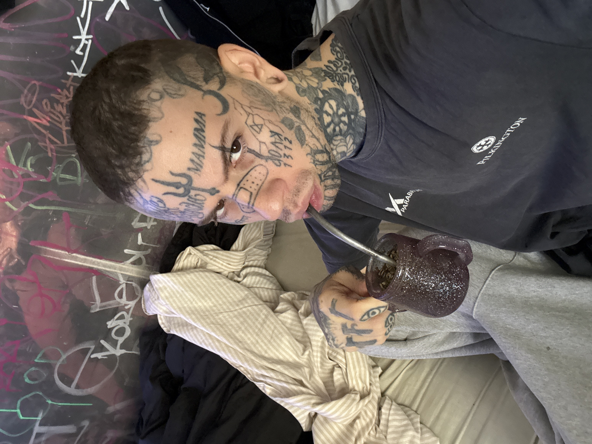 An intimate, angled portrait captures a young adult male with distinctive features and extensive body art. He gazes directly at the camera with a calm expression. His face, neck, and hands are heavily tattooed; notable facial tattoos include "MAMMA" on his forehead, a band-aid graphic on his right cheek, a trident symbol near his left eye, and the letters "KAVN" followed by "312" on his right cheek. Elaborate floral and geometric designs cover his neck and the visible parts of his arms and hands, with an eye symbol on his fingers.
The man is actively drinking from a glittery purple mate gourd through a metal bombilla, which is positioned in his mouth. The gourd contains yerba mate. He wears a dark t-shirt featuring a white "PILKINGTON" logo and a partially visible "PARABR" logo. His lower body is clad in casual grey trousers or sweatpants.
The setting appears to be an indoor, informal space, possibly a home. In the background, a dark, reflective surface is covered in various graffiti-style markings in white, pink, and green, with a peace sign and the letter "K" being discernible. Discarded or rumpled clothes, including a striped beige and white fabric and a black item, are visible around him. The consumption of mate, a prominent cultural practice, aligns with the given location context of Buenos Aires, Argentina.