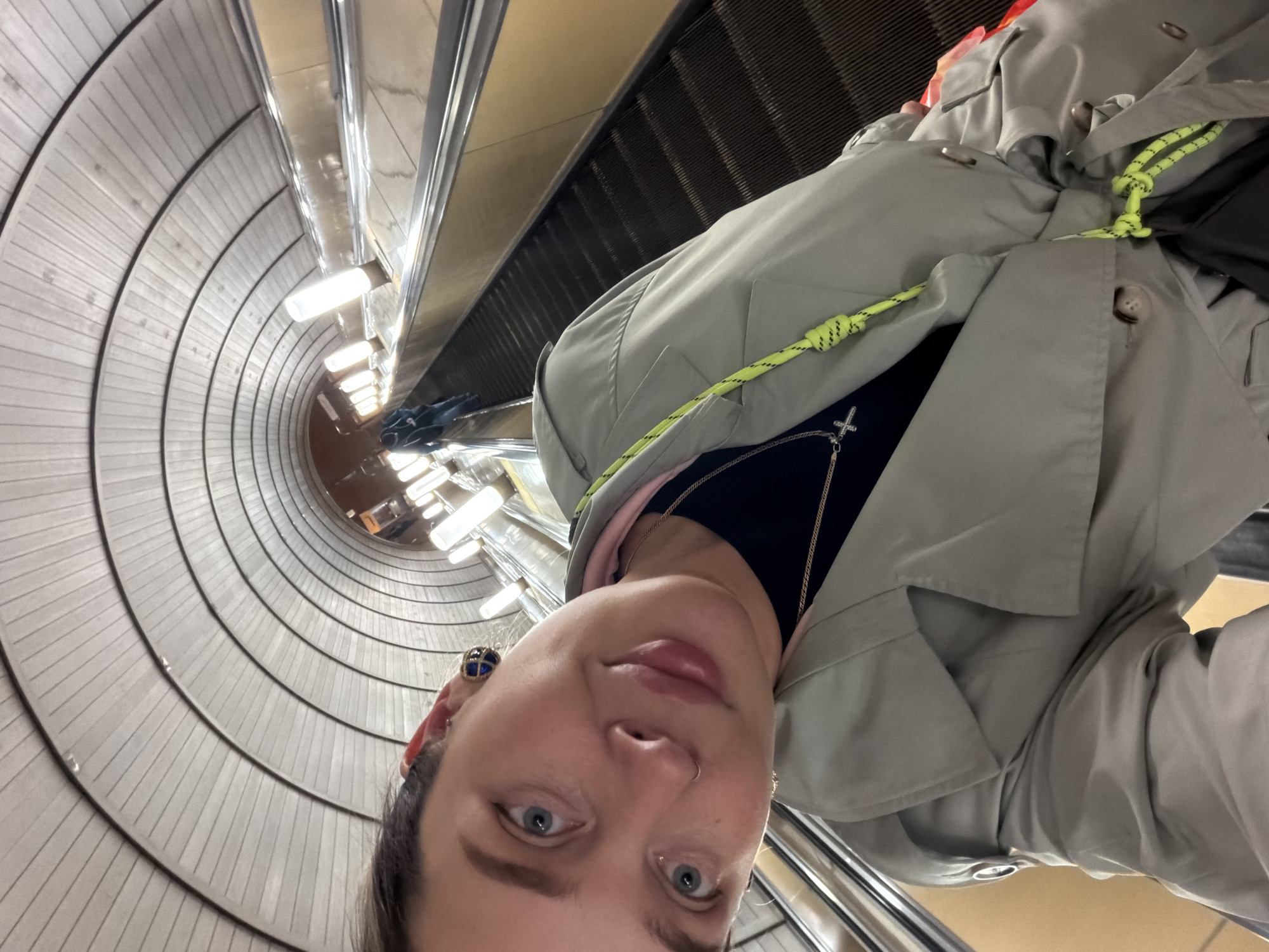 A selfie photograph taken inside the Moscow Metro in Russia shows a woman looking into the camera while standing on an escalator. The escalator ascends a long, cylindrical shaft lined with evenly spaced, horizontal ribs and illuminated by evenly spaced linear light fixtures. The woman's face is framed by the tunnel. She has light skin, blue eyes, and dark hair and is wearing an earring with a blue stone, a navy blue top, and a light gray jacket with a fluorescent yellow drawstring. She is also wearing a silver chain necklace with a cross pendant. The perspective indicates that the photo was taken upwards from the escalator.