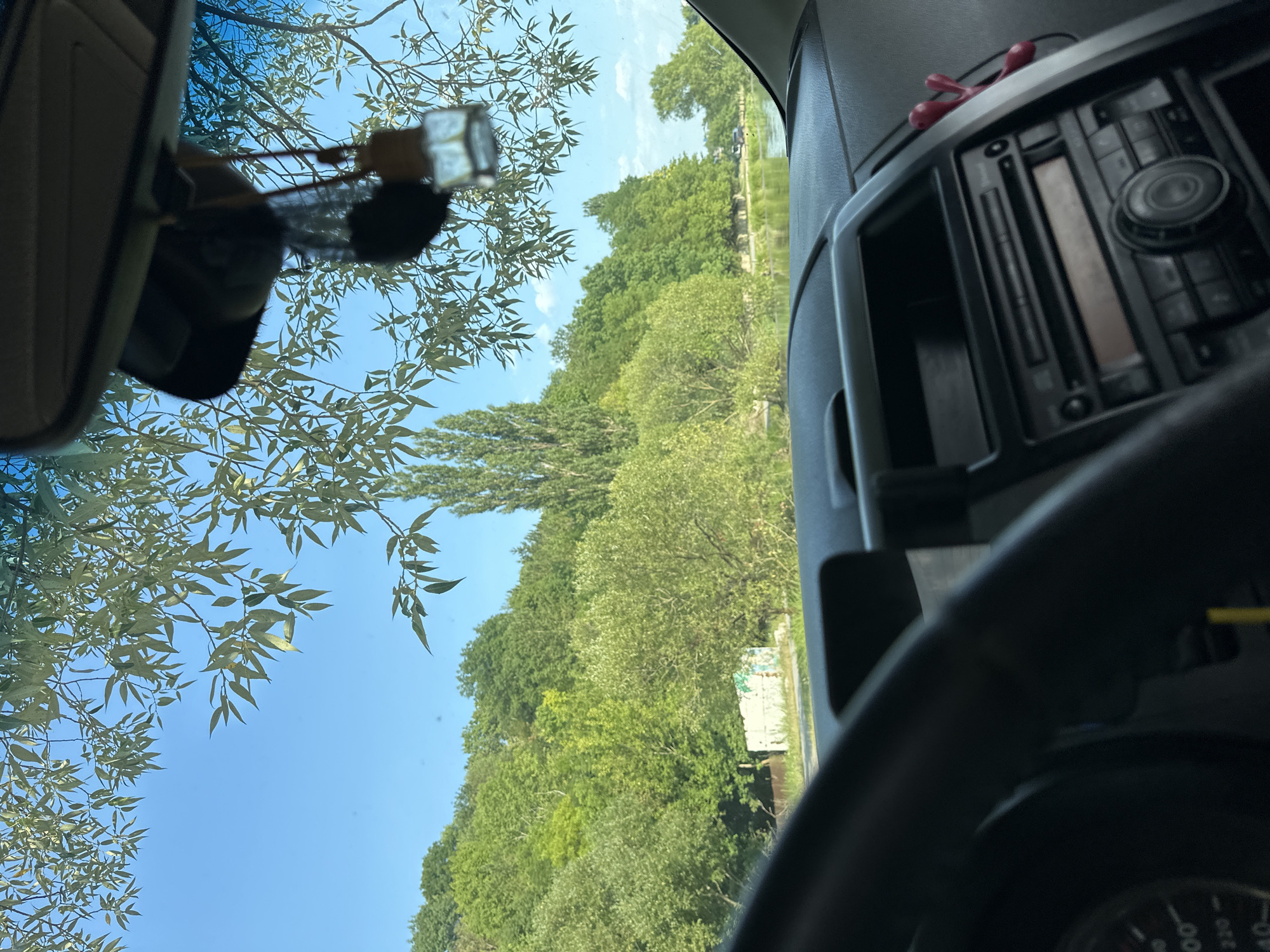 The image, taken from inside a car, shows a view through the windshield on a clear, sunny day. In the foreground, the dashboard and steering wheel are visible, with a car's audio system and a dangling air freshener. Outside, a lush green hillside covered in trees dominates the landscape, with a body of water, possibly a river or lake, in the middle ground. The sky is a vibrant blue, with some tree branches and leaves framing the top of the scene. The location is stated to be Makiv, Ukraine.