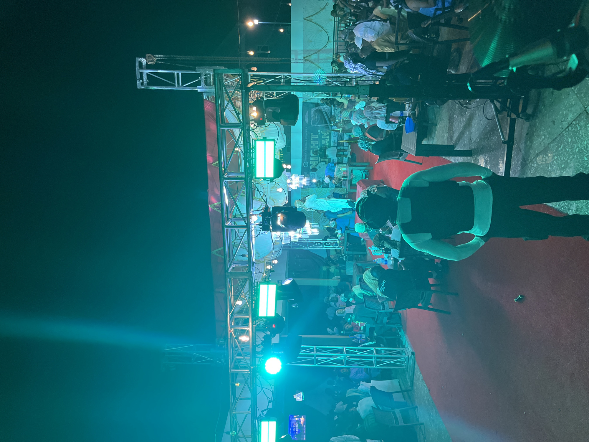 The image depicts an outdoor or semi-outdoor event at night, dominated by a teal/green artificial light cast, in the city of Unknown, Unknown.
In the immediate foreground, a person stands with their back to the viewer, slightly right of center, wearing dark trousers, a light-colored long-sleeved shirt, and a black vest, with a dark head covering or headphones. They are positioned on a multi-toned concrete surface. To their left, a red carpet extends from the mid-ground into the foreground. On the red carpet, two small, green cylindrical objects lie on the ground.
The mid-ground features a seated audience of approximately 50-70 individuals at utilitarian dark tables and chairs. Many male attendees wear traditional headscarves or hats, and multiple women wear head coverings. Beverages in bottles are visible on tables. To the right, partially obscured, is a drum set or musical equipment.
The stage area, located in the mid-ground to background, is framed by a metal truss system supporting various lights, including a prominent circular green spotlight on the left and rectangular green lights centrally. String lights with warm yellow bulbs are strung across the top truss. A person in white robes stands on the raised stage, facing the audience, appearing to perform or speak. Another individual is visible near them, also on stage. A banner on the stage backdrop reads "SHIKAD GLOBAL CONCEPT" and "NIGHT LAN" (partially visible). Speakers are mounted beneath the stage. Decorative chandeliers hang from the truss system above the audience.
A tall metal truss structure stands on the far right, from which a metal hook and chain are suspended. In the upper left, a strong, narrow beam of teal light projects upwards into the dark night sky. The overall scene is brightly lit by the artificial teal/green lighting, creating strong shadows and obscuring precise details in some areas.