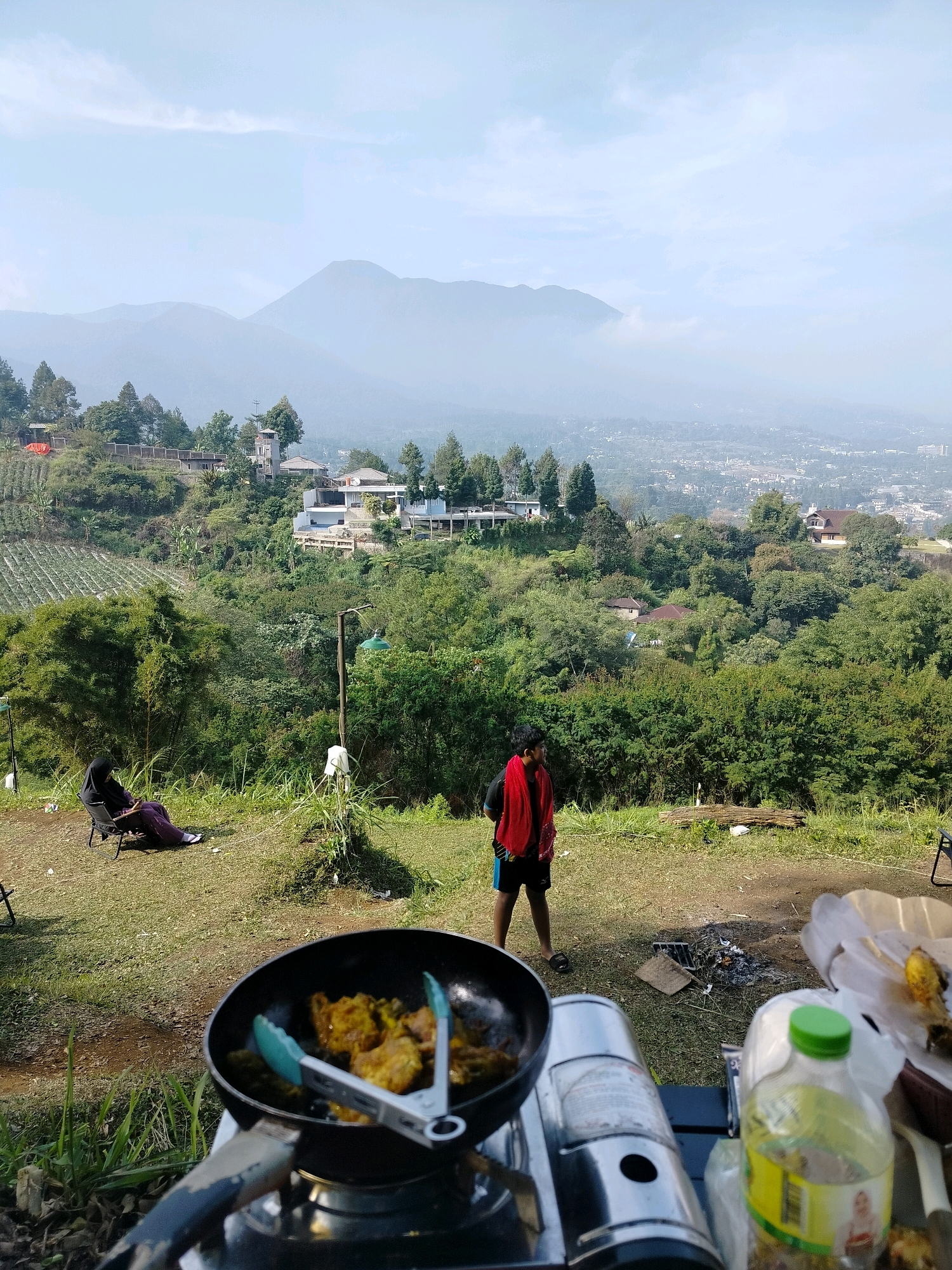 The image depicts a scenic outdoor setting with a view of rolling hills and a distant city. In the foreground, a portable gas stove is set up with a frying pan containing what appears to be fried food. Tongs are placed inside the pan. To the right of the stove, a partially unwrapped plastic bag with food and a green-capped plastic bottle are visible.
Further back, a young person wearing a red scarf around their neck and dark shorts stands facing away from the camera, looking towards the landscape. To the left, a person in a black hijab and purple clothing sits in a folding chair.
The environment consists of grassy terrain with scattered trees and vegetation. In the middle ground, there are more trees and some buildings, suggesting a residential area or resort on the hillside. The background is dominated by hazy mountains and a view of a city nestled in the valley. The sky is bright with some clouds. The overall atmosphere suggests a leisurely outdoor activity, possibly a picnic or camping trip, with a cook preparing food. The presence of buildings and the specific terrain are consistent with the hilly landscape of Cisarua, Indonesia, known for its natural beauty and vacation spots.