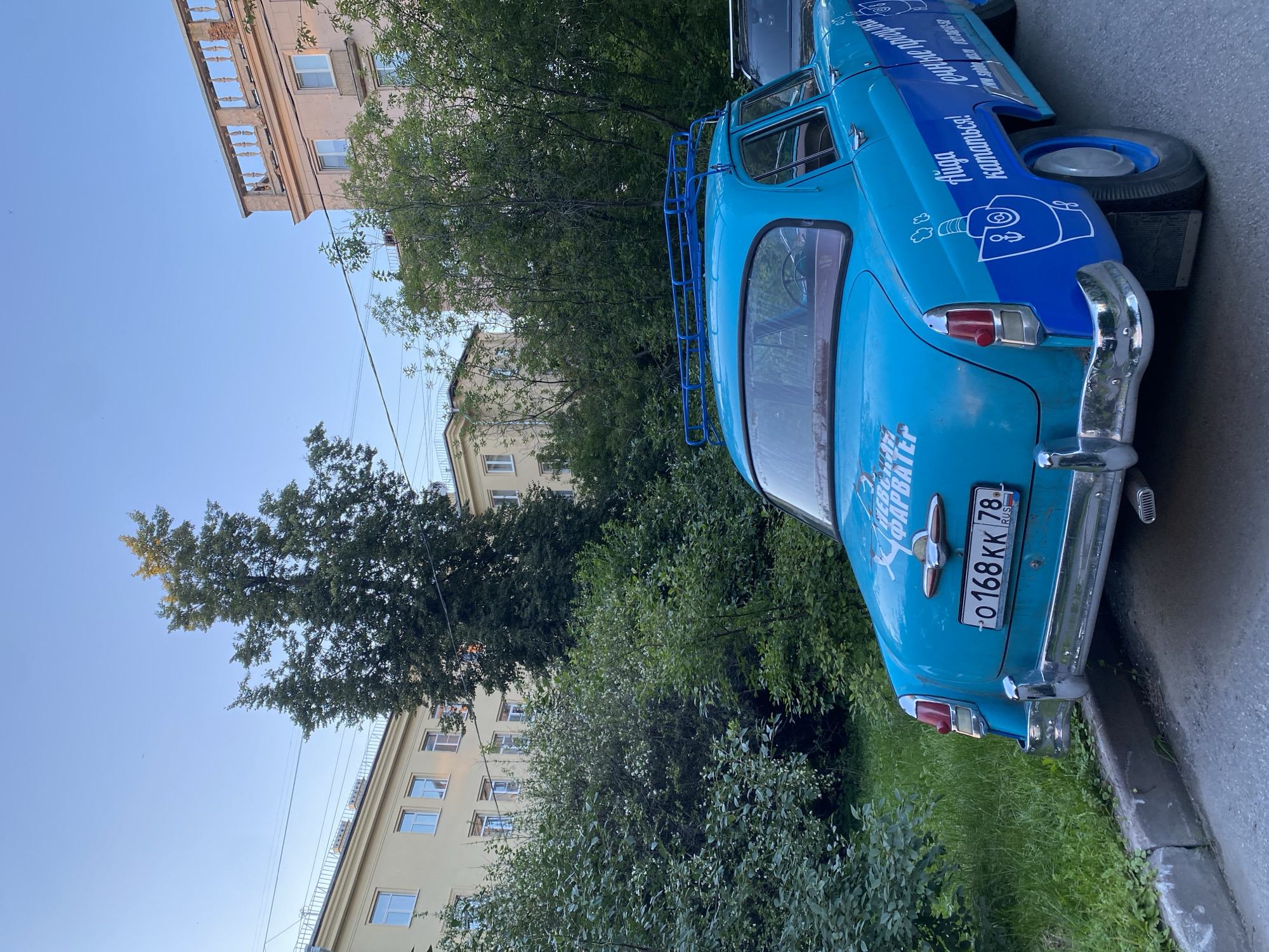 The image captures the rear and side view of a vintage, bright blue sedan parked on a street. The car features a blue roof rack and decals on its sides and trunk. One decal on the trunk reads "О168КК 78 RUS" which is a Russian license plate number. Additional decals on the side of the car include an illustration of a ship and text that appears to be in Cyrillic, possibly promotional text. The car is parked alongside a grassy area and a concrete curb, with trees and apartment buildings visible in the background. The buildings are multi-story, with windows and balconies, and appear to be located in an urban setting. The sky is clear and blue, suggesting daylight. The presence of the "78" on the license plate indicates the vehicle is registered in Saint Petersburg, Russia.