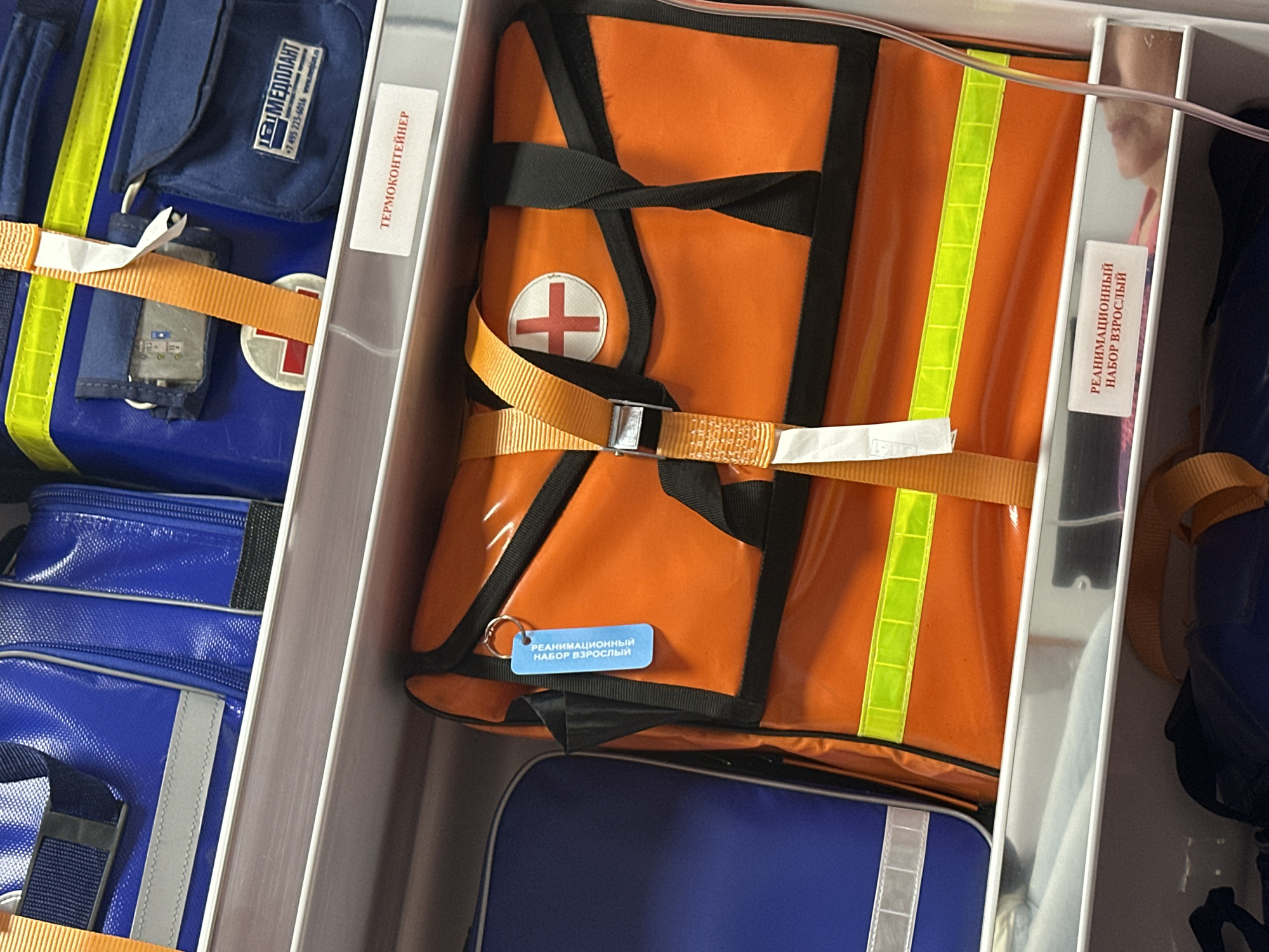 The image shows the inside of a storage compartment, possibly in a medical or emergency response setting. Visible are several medical bags and containers. One bag is bright orange with black straps, a white circle containing a red cross, and a reflective yellow stripe. A small blue tag is attached to the bag with the Russian text "РЕАНИМАЦИОННЫЙ НАБОР ВЗРОСЛЫЙ," which translates to "RESUSCITATION KIT ADULT." Other containers are blue. The text on one of the containers is "ТЕРМОКОНТЕЙНЕР," which translates to "THERMO CONTAINER." The label on a blue bag says "МЕДПЛАНТ" ("MEDPLANT") and a phone number with the country code +7, indicating it is likely in Russia. The location is not discernable from this image, but the Medplant location is Moscow.