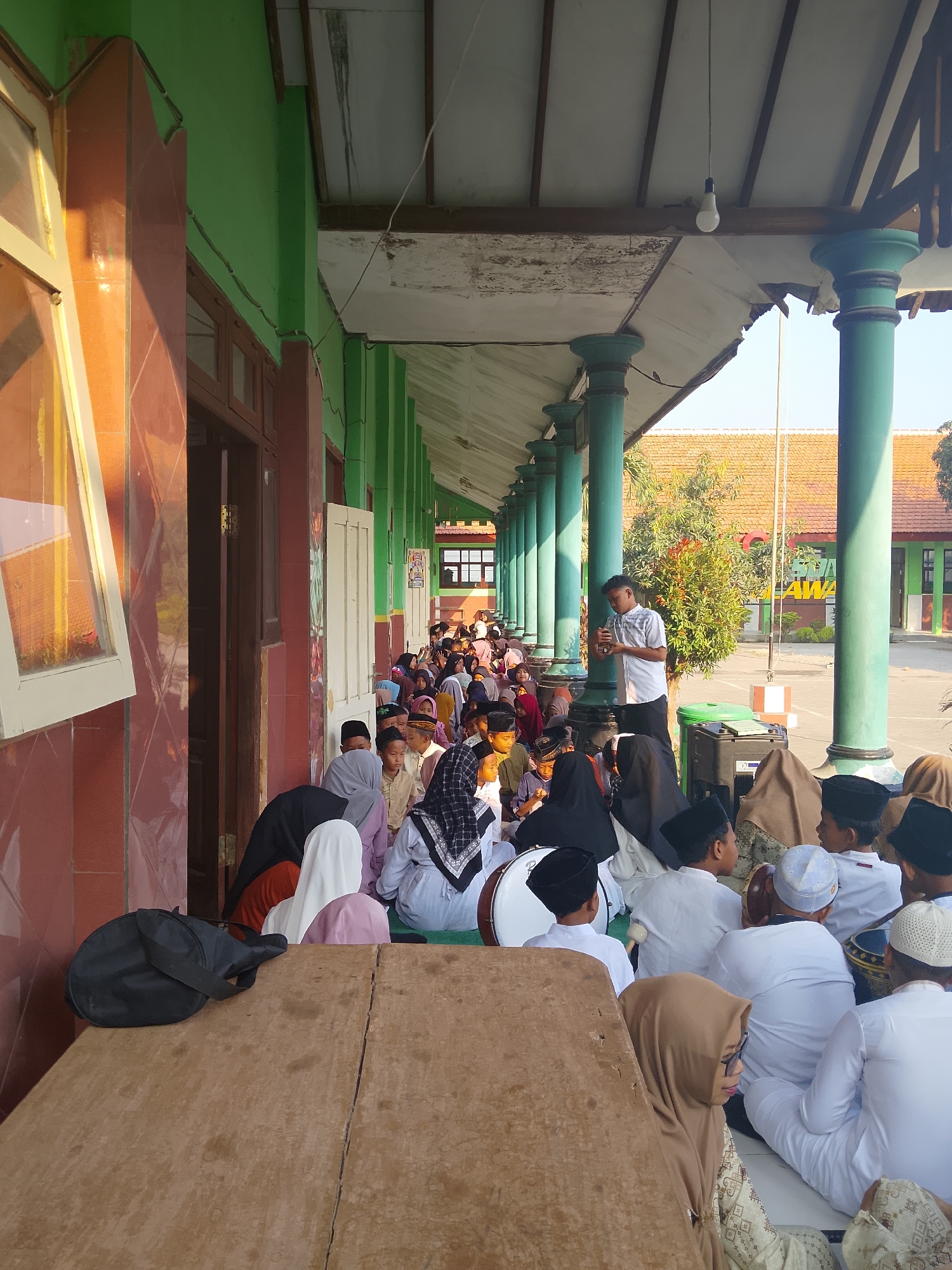 The image depicts an outdoor gathering of people, likely students, in a covered walkway or veranda of a building with green painted walls and pillars. The location is identified as Kepuh, Indonesia. A significant number of individuals, predominantly children and young adults, are seated on the ground, facing a young man who is standing and holding a microphone, appearing to address the group. Many of the attendees are wearing Islamic attire, including peci hats and hijabs. In the foreground, a wooden surface, possibly a table or platform, is visible with a black bag placed on it. To the right of the standing speaker, a black audio speaker is positioned. In the background, other buildings with orange tiled roofs and trees are seen under a clear sky. The overall atmosphere suggests an organized event such as a school assembly or religious gathering.