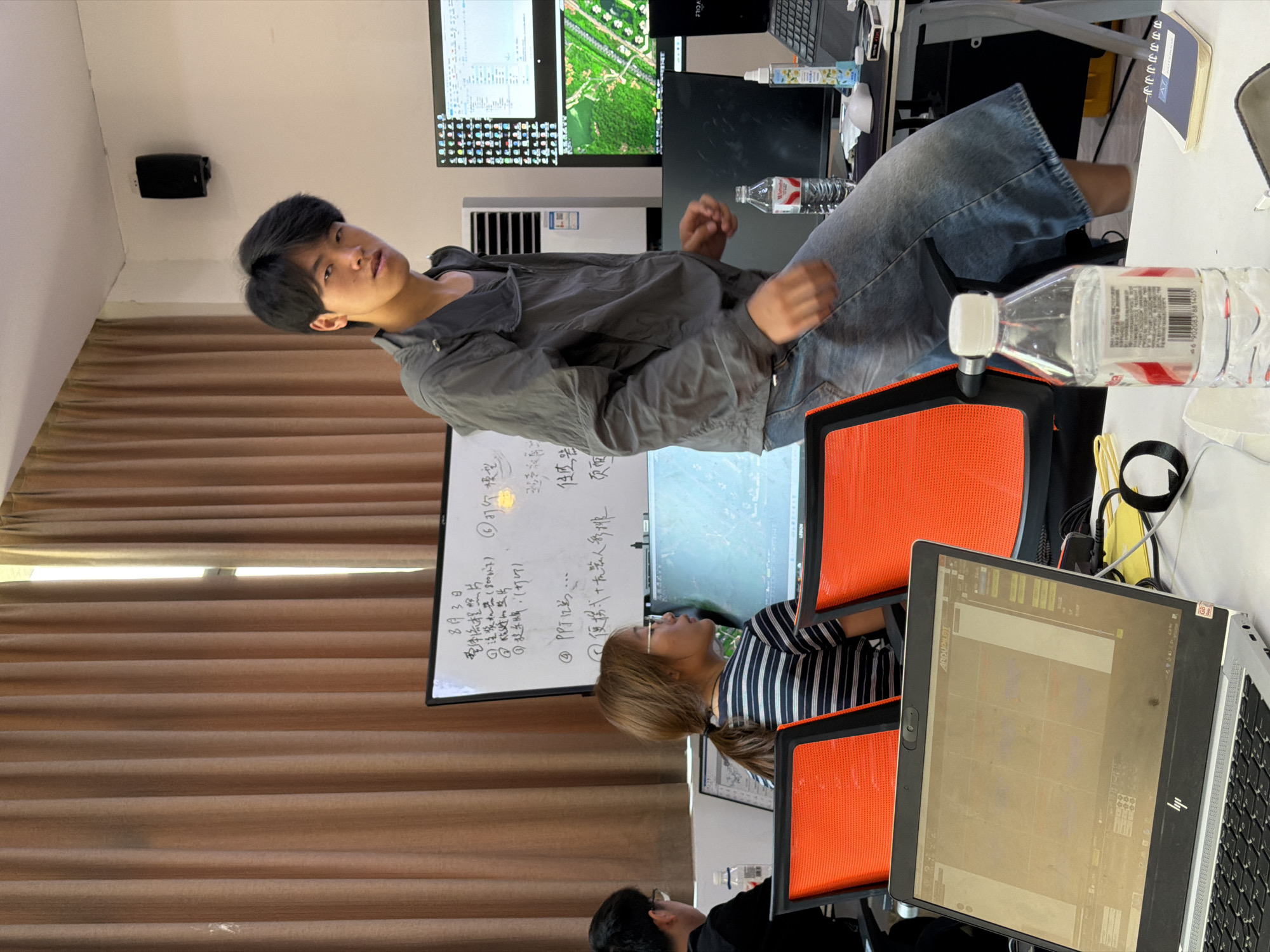 The image depicts an indoor scene, possibly an office or classroom, with a light brown curtain in the background. On a whiteboard behind the individuals, Chinese characters are visible, suggesting it's a location in China, specifically Jiangjin. In the foreground, a young man in a grey jacket and denim shorts is standing, looking towards the camera. To his left, a woman with her hair in a ponytail, wearing a striped shirt and glasses, is seated and looking at a computer monitor. A laptop is open on a desk in front of her. Other computer monitors and office furniture are visible, including bright orange chairs. A bottle of water is placed on the desk near the young man.