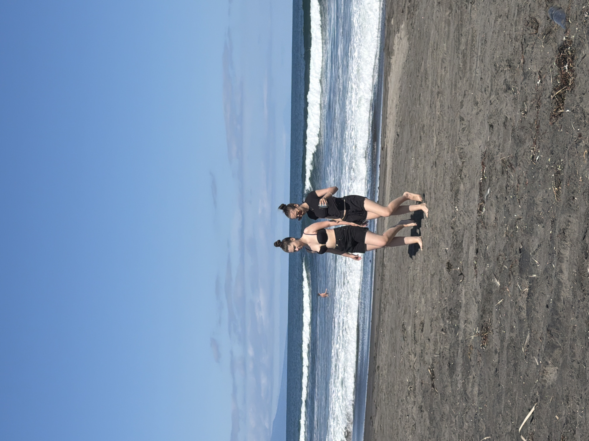 Two women are walking on a dark sand beach in Yuzhno-Kurilsk, Russia. The woman on the left is wearing a black bikini top and black shorts, and the woman on the right is wearing a black t-shirt and black shorts. They are walking towards the right side of the frame. In the background, there are waves crashing on the shore, and a person can be seen in the water. The sky is blue with some clouds. The overall environment is a coastal scene on a sunny day.