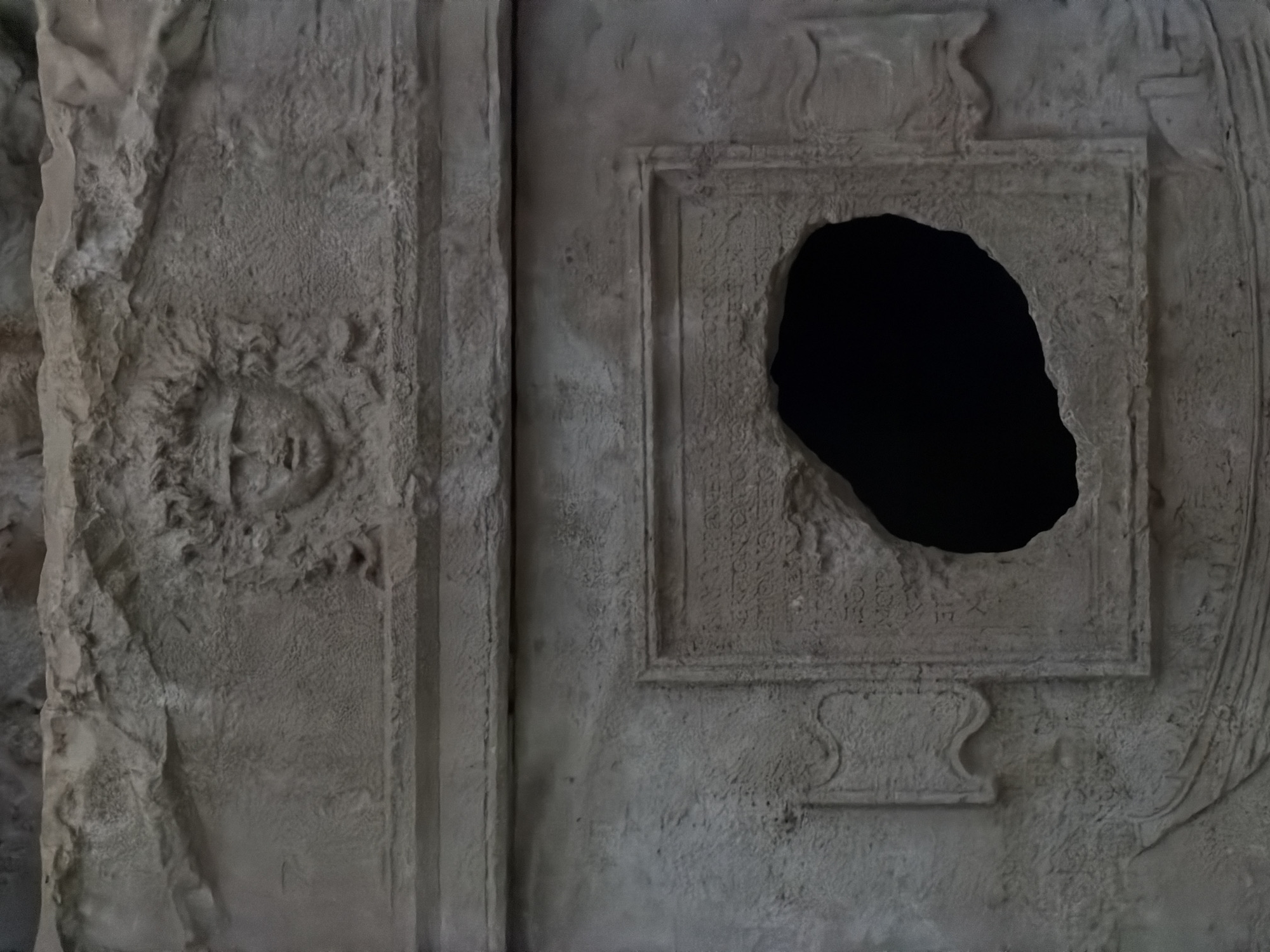 The image displays a close-up of an ancient, weathered stone surface, likely part of a tomb or monumental structure in Kumluca, Turkey. The central feature is a large, irregularly shaped, deep black void or hole within a rectangular, framed panel. This panel, carved into the stone, shows faint, eroded inscriptions to the left of the opening, which appear to be an ancient script, possibly Lycian or Greek, with several lines of characters visible. Below this main panel, decorative S-shaped curves, resembling volutes or handles, are carved into a lower section.
To the left of the central panel, a heavily eroded circular relief is discernible. This motif appears to be a stylized head or sun symbol, with a very faint, indistinct face-like feature at its center surrounded by radiating or hair-like patterns. A vertical, column-like band separates this left section from the central framed panel. The entire stone surface exhibits significant age and weathering, with rough textures and areas of erosion. The dim, diffused lighting enhances the sense of antiquity and mystery, suggesting an entrance into a dark chamber or a damaged section of a historical site.