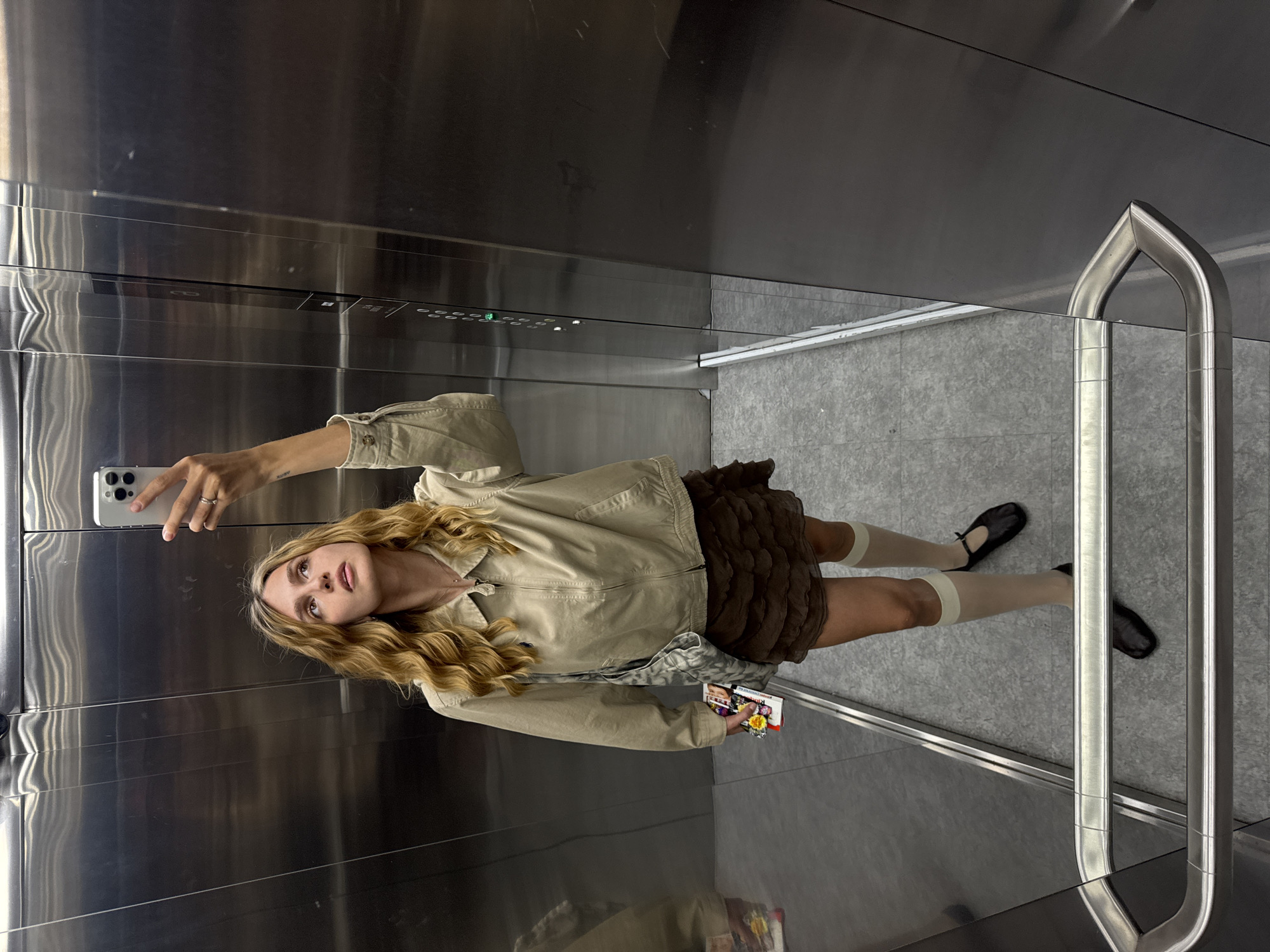 A young woman stands in an elevator, taking a selfie with her smartphone. The elevator's interior is stainless steel with a grey floor and a vertical handrail on one side. She is wearing a khaki jacket, a brown ruffled skirt, beige knee-high socks, and black ballet flats. She holds a small book or magazine in her other hand. The elevator buttons are visible on the back wall. The location is Unknown, Unknown.