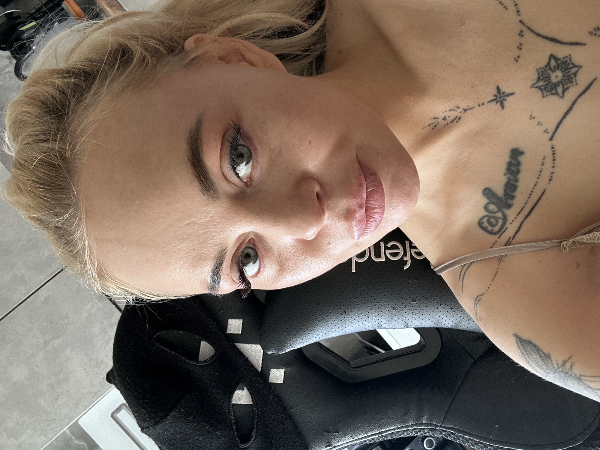 A medium close-up, high-angle selfie depicts a woman in an indoor setting in Unknown, Unknown. She is fair-skinned with blonde hair, pulled back, and her face is positioned slightly below the center of the frame, looking directly at the camera. Her eyes are light green/hazel, with visible dark eyelashes (possibly false) on both lids. Her eyebrows are defined and dark. Her lips are full, slightly parted, and show a light pink tint.
The woman wears a light beige or grey camisole with a thin strap visible on her right shoulder (viewer's left). Multiple black line-work tattoos are visible on her chest and clavicle area, including a script-style tattoo on the left side (partially legible as "@Annian") and a starburst-like geometric design below it. Smaller dotted and linear patterns are also present. A larger, lighter-ink tattoo, possibly a feather, is visible on her left upper arm/shoulder.
To the left and slightly behind her is a black gaming or office chair made of perforated leather-like material, with the word "defend" printed vertically on its side. Draped over the chair's headrest is a black fabric object, possibly a cap or balaclava, with two white eye-like cutouts and small white square patterns. The background in the upper left corner shows a light grey, possibly tiled, wall surface, with a thin white horizontal element. The upper right background is darker and blurred.