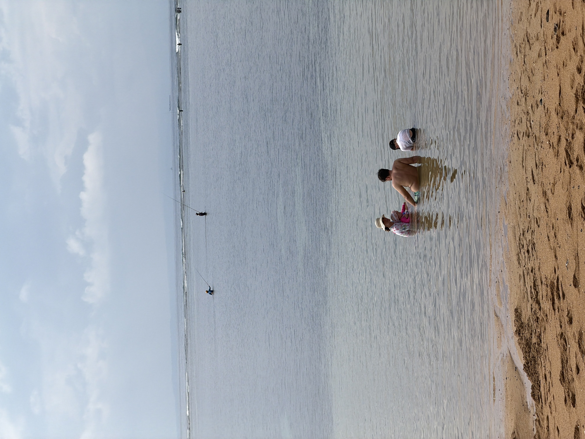 A serene coastal scene unfolds in Denpasar, Indonesia, with a sandy beach in the foreground giving way to calm, shallow ocean waters. In the middle ground, a family enjoys the water: an adult male, shirtless, is partially submerged, with a young child in a pink bathing suit and a straw hat to his left, and another child in a white t-shirt to his right. Further out in the ocean, several individuals are visible, some holding fishing rods, suggesting a popular fishing spot. The horizon is marked by a hazy outline of land and a faint ship in the distance. The sky is overcast with soft, diffused clouds, creating a muted light over the seascape. The overall atmosphere is one of peaceful outdoor recreation.