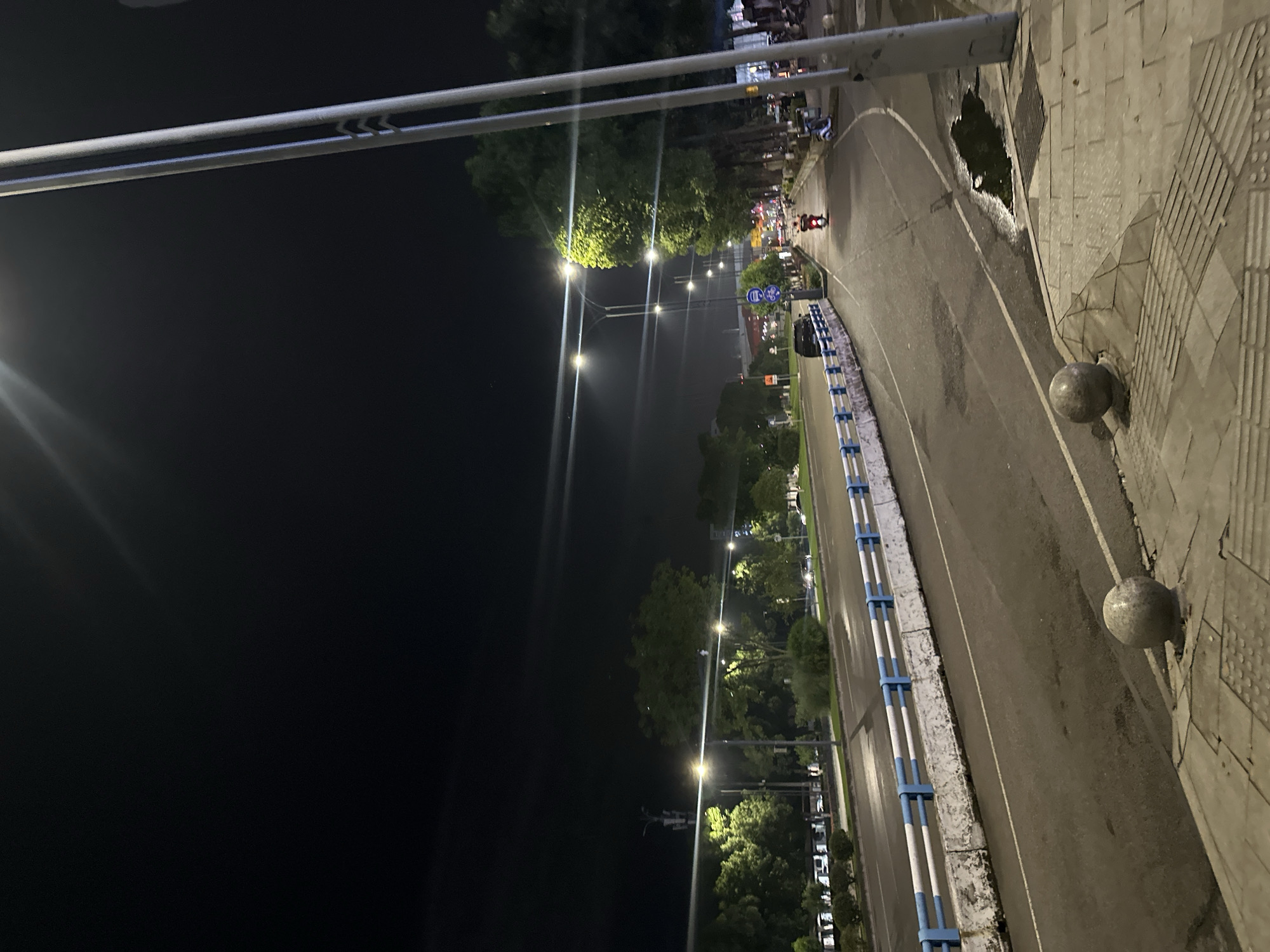The image depicts a nighttime scene on a street in the Chongqing High-tech Zone, China. A two-lane road curves to the right, illuminated by streetlights that cast long shadows. On the left side of the road, a blue and white barrier separates the roadway from a sidewalk area lined with trees and bushes. Several cars are visible on the road, and a motorcycle is moving away from the camera on the right side of the road. A tall lamppost stands prominently in the foreground on the right, with another visible further down the street. The ground appears wet in places, suggesting recent rain. The sky is dark and clear, with no visible stars.