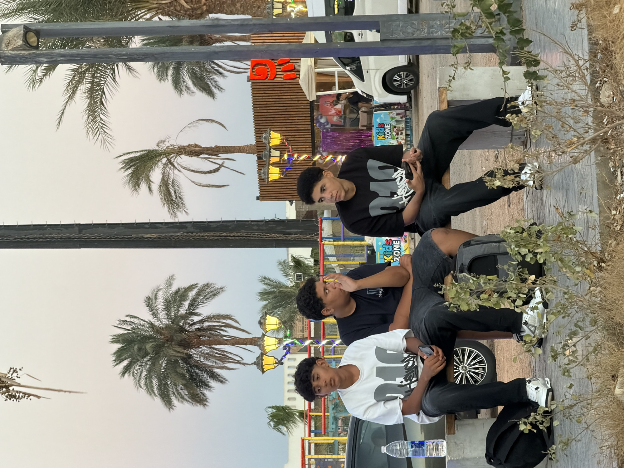 Three young men are seated on a bench in what appears to be an outdoor public area, possibly a promenade or entertainment district in Sharm El Sheikh, Egypt. The man on the far left is wearing a white t-shirt with gray graphic text, black pants, and white sneakers. He is holding a mobile phone. The man in the middle is wearing a black t-shirt with graphic text, dark shorts, and is looking to his right with his hand near his face. The man on the far right is wearing a black t-shirt with graphic text, black pants, and white sneakers. He is holding a mobile phone and looking to his left. Two black backpacks are placed on the ground next to the bench. A bottle of water is on the bench near the man on the left. In the background, palm trees are visible, along with decorative lights and some structures that suggest a commercial or entertainment venue. A white van is parked behind the bench. The overall lighting suggests it is either early morning or late evening.