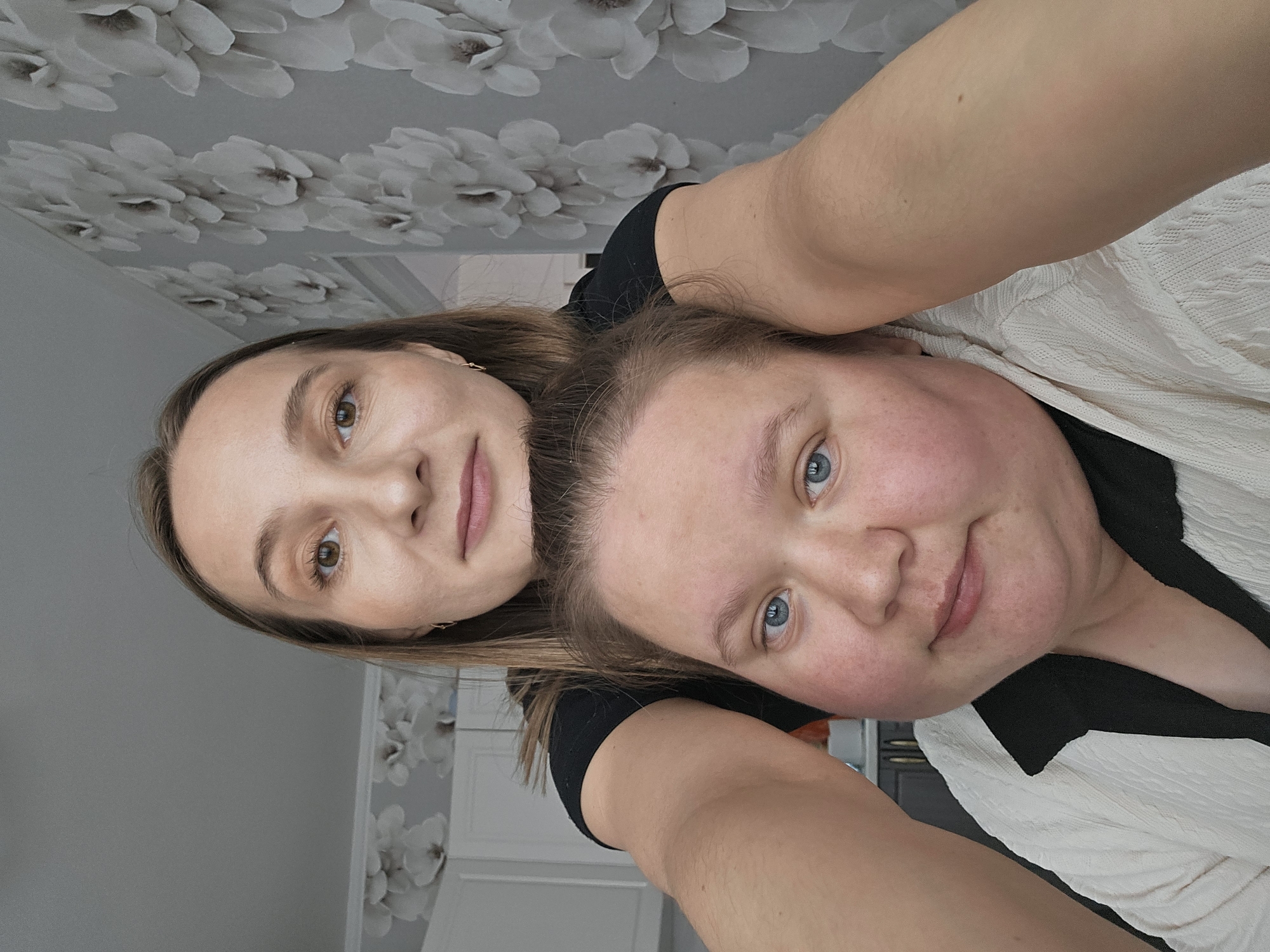 Two women are posing for a selfie in an indoor location with floral wallpaper. The woman on the left has brown hair and brown eyes. The woman on the right has lighter brown hair and blue eyes. The location appears to be in the city of Chaikovsky, Russia.
