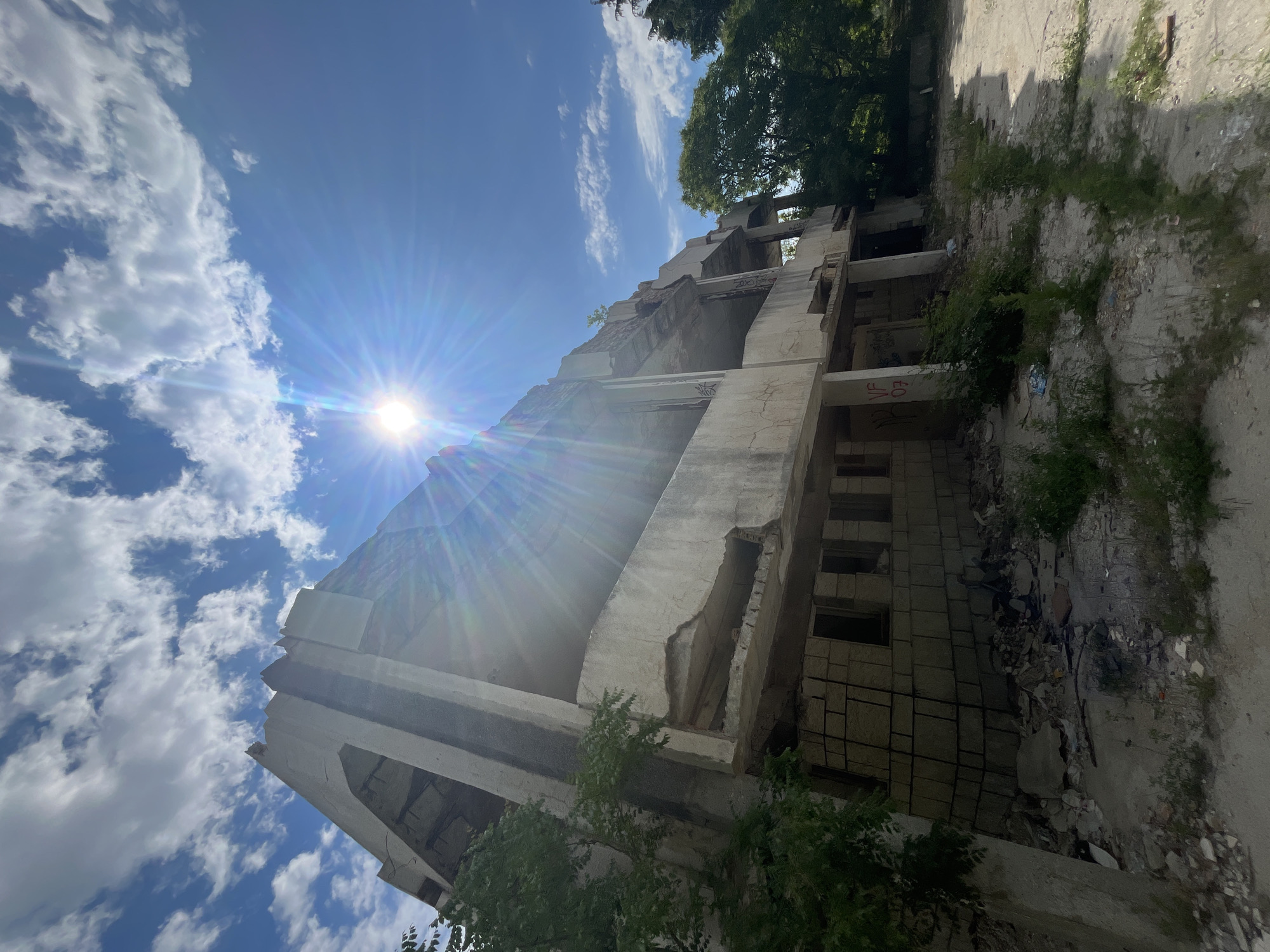 The image captures an abandoned, multi-story concrete building bathed in bright sunlight. The structure shows signs of significant decay, with crumbling concrete walls, exposed rebar, and broken window openings. The building's facade is weathered, and graffiti is visible on some sections. Overgrown vegetation, including trees and weeds, encroaches on the building and the surrounding unpaved ground, which is littered with debris. The sky above is a vibrant blue, dotted with fluffy white clouds, and the sun's glare creates a starburst effect, casting lens flare across the scene. The overall impression is one of neglect and disrepair. The building is located in the city of Varna, Bulgaria.