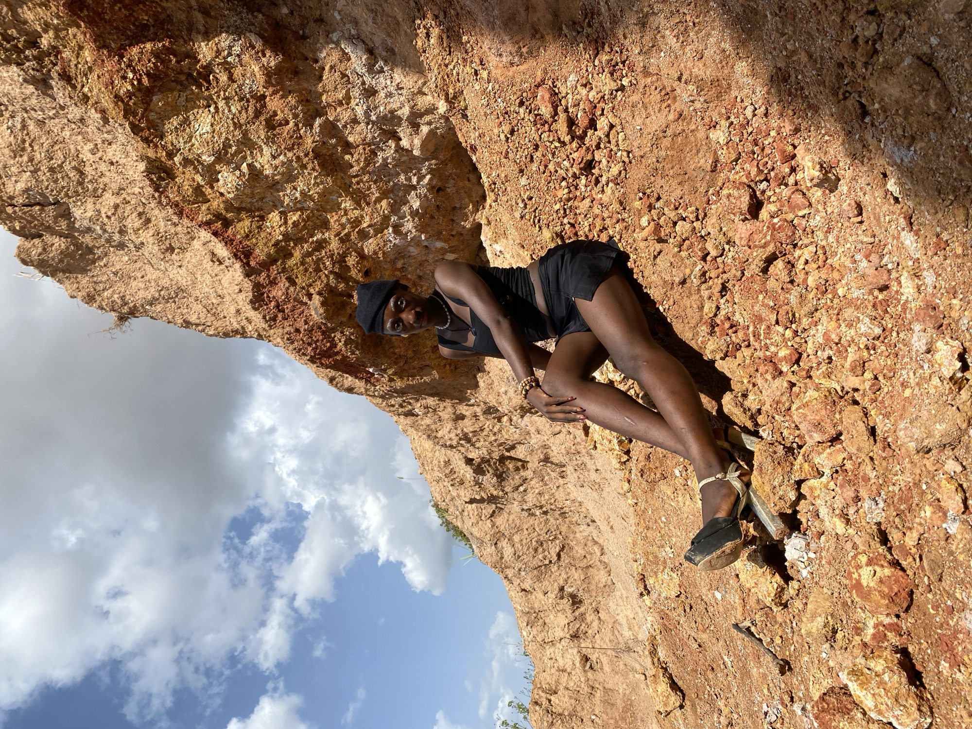 A person is posing on a rocky, reddish-brown cliff face under a bright, cloudy sky. The person, wearing a black beret, a black top with thin straps, and black shorts, is lying down with their legs bent and crossed. They have on black high-heeled sandals with ankle straps and are adorned with a pearl necklace and a beaded bracelet. The setting appears to be outdoors in a dry, arid environment with exposed rock and dirt. The lighting suggests it is daytime, possibly midday, with strong sunlight casting shadows on the rocks. The sky is partly cloudy with patches of blue visible. The image is oriented vertically, with the person positioned in the lower right quadrant of the frame, leaning against the rugged terrain. The overall mood is relaxed and fashionable, with the subject striking a pose against the natural backdrop.