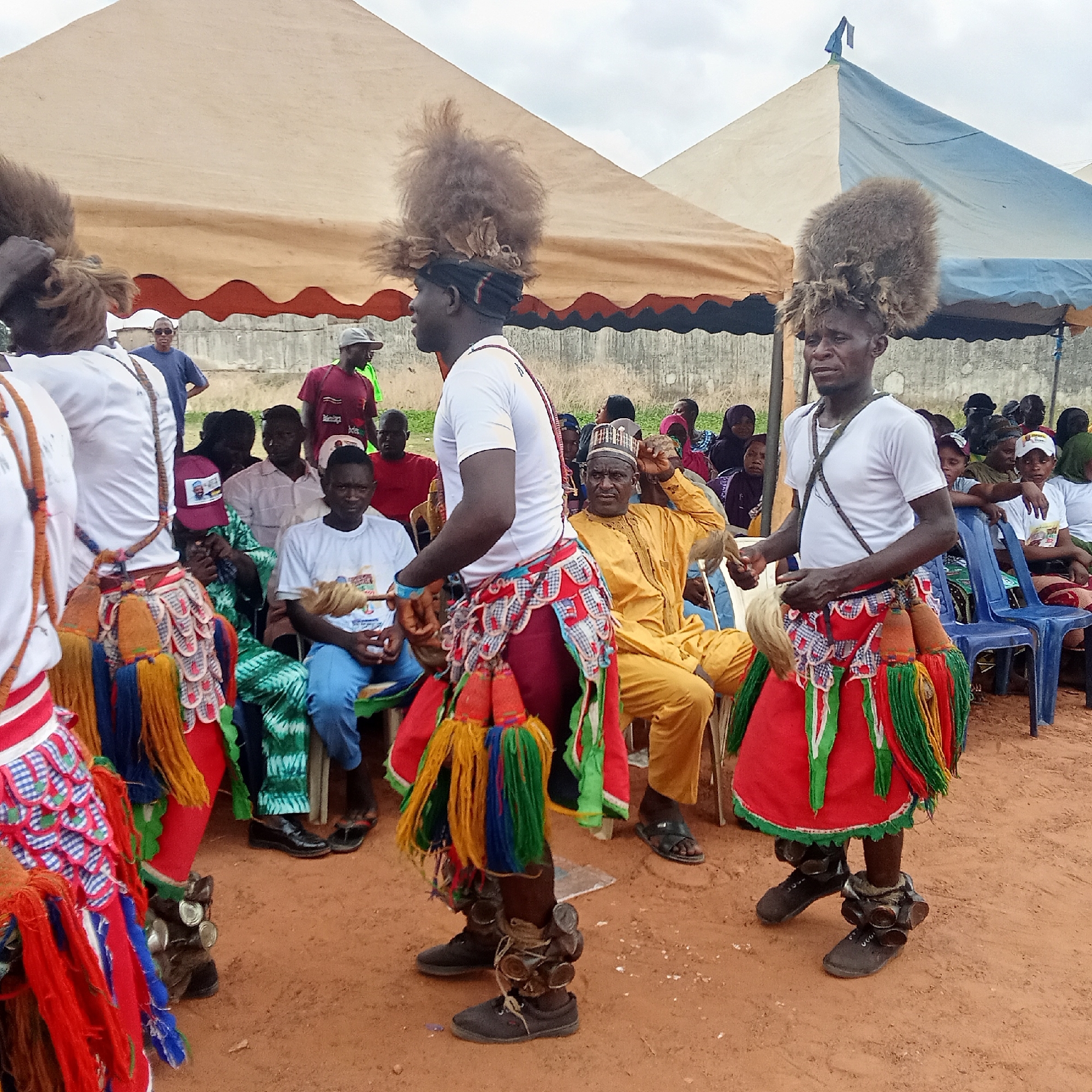 Akwanga, Nigeria: several men in traditional attire dance for a seated audience. The dancers wear white t-shirts, elaborate skirts with tassels, and tall headdresses made of fuzzy material. Metallic objects are tied to their shoes. An audience watches from chairs set up under two tents. The ground is reddish-brown.