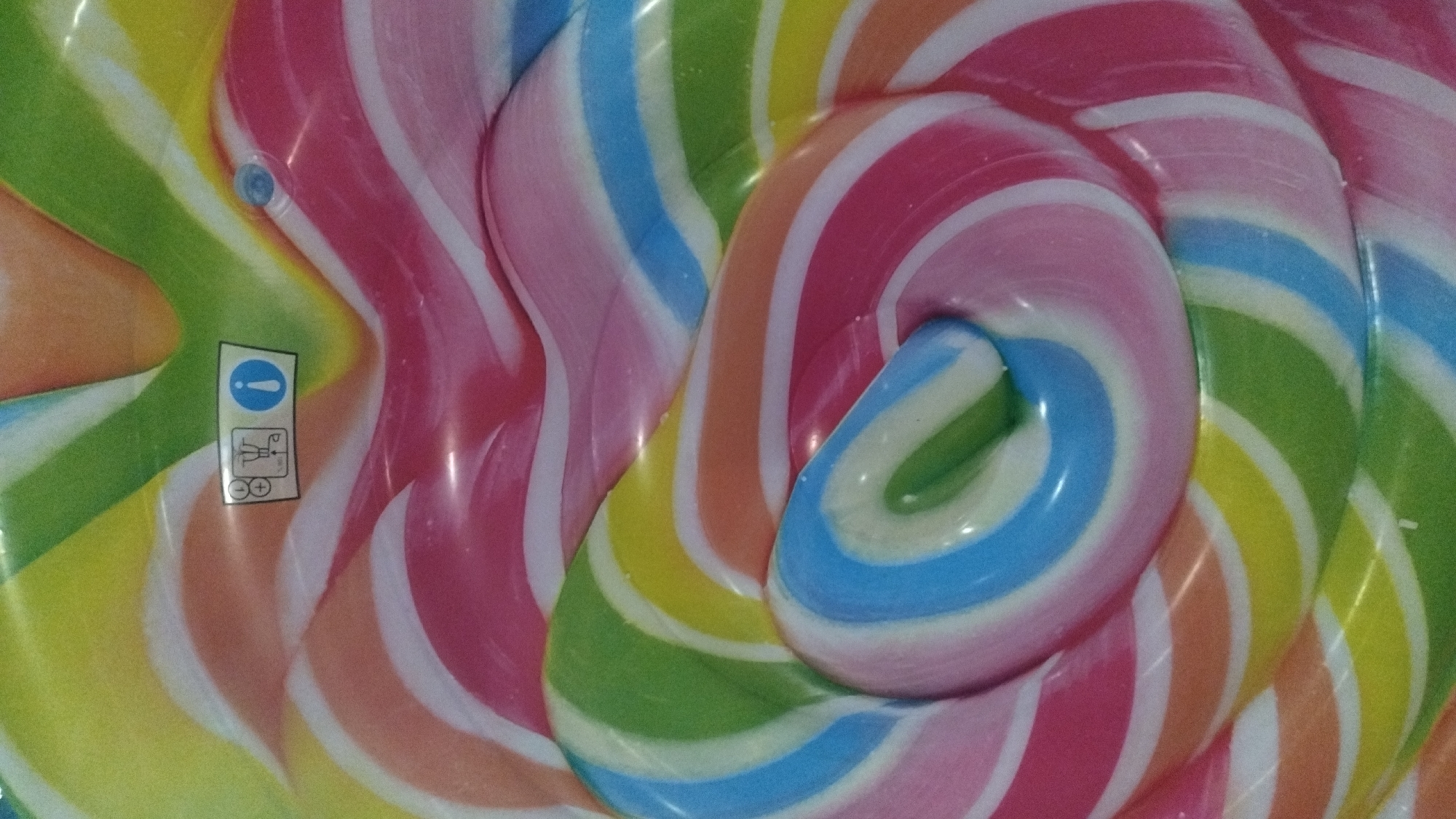 The image is a close-up, abstract shot of a colorful, swirling pattern resembling a giant lollipop. The colors are vibrant and include pink, orange, yellow, green, light blue, and white, arranged in curved stripes. There is a small, rectangular label with icons on it, positioned in the upper middle of the frame. To the right of the label, a small blue inflation valve is visible. The surface appears to be smooth and possibly inflatable, given the valve. There are no people or any recognizable landmarks of Perea, Greece present in the image. The scene is a close-up of a manufactured item, devoid of any environmental context that would link it to a specific location like Perea.