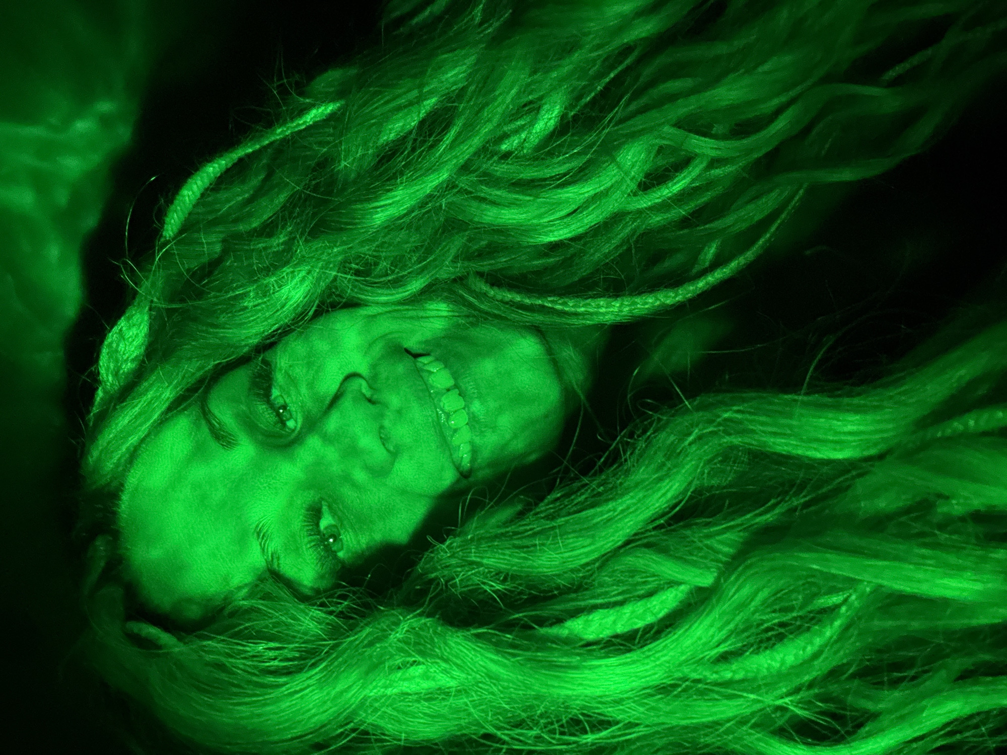 A close-up, low-angle photograph depicts a person lying down with long, green hair spread around their face. The person's face is illuminated by a green light source, casting a vibrant green hue over the skin and hair. The individual is smiling, with teeth visible. The background is dark and undefined. The image shows the face of a girl laying on the floor in green light with hair.
This image does not contain information to suggest that this image was taken in Rusanovo, Russia.