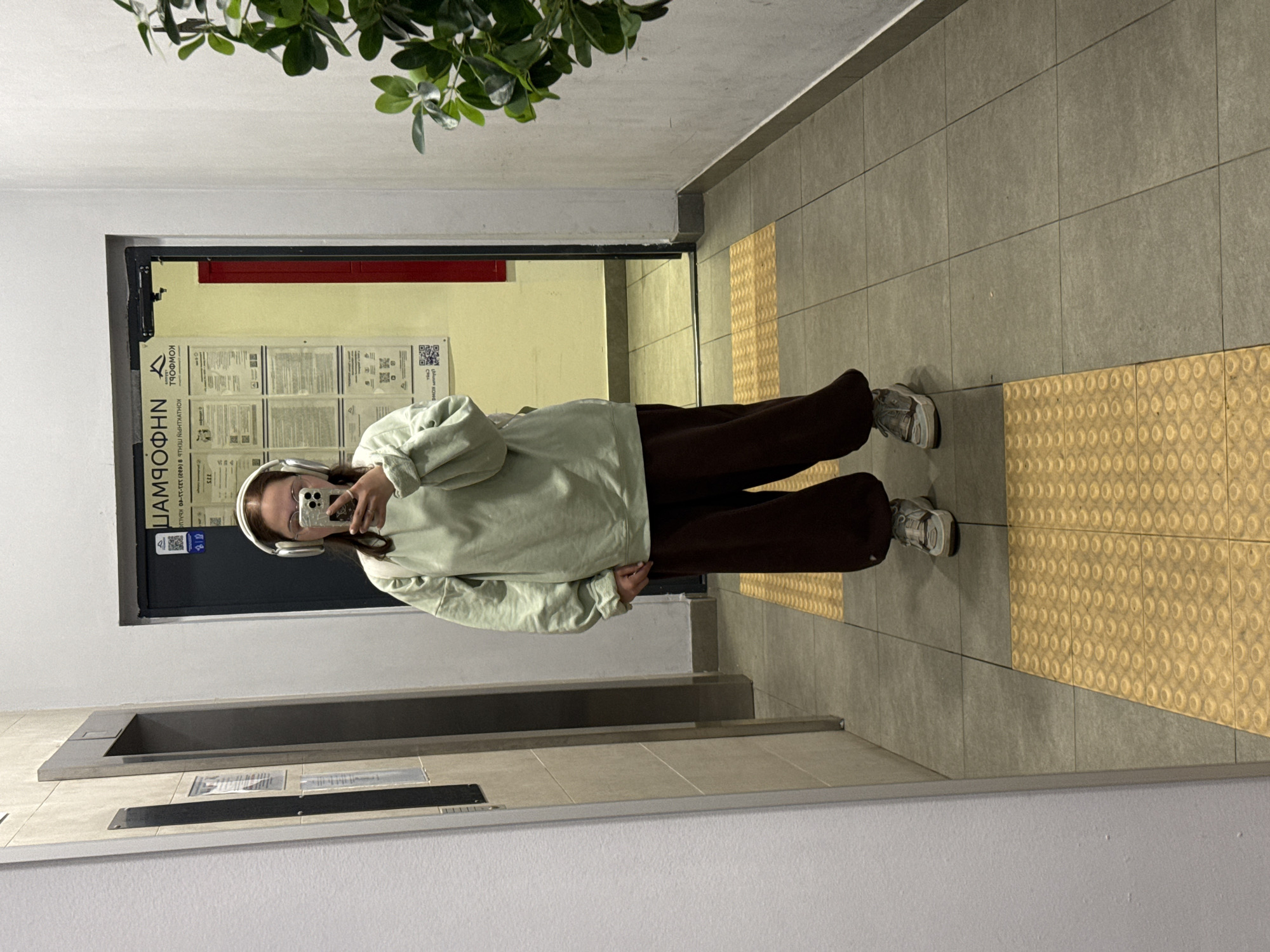 A full-body, front-facing mirror selfie taken indoors shows a young woman wearing a light green oversized sweatshirt, dark brown baggy sweatpants, and gray sneakers. She has on white headphones and is holding a smartphone in her right hand, angled to capture the reflection. The background reveals an interior hallway, possibly in a residential building, with an elevator door to the left. A bulletin board with text and QR codes is visible on the wall behind her, and a plant with green leaves is on the right side of the frame. The floor is tiled with a tactile paving strip in yellow. The location is identified as Lyubertsy, Russia.