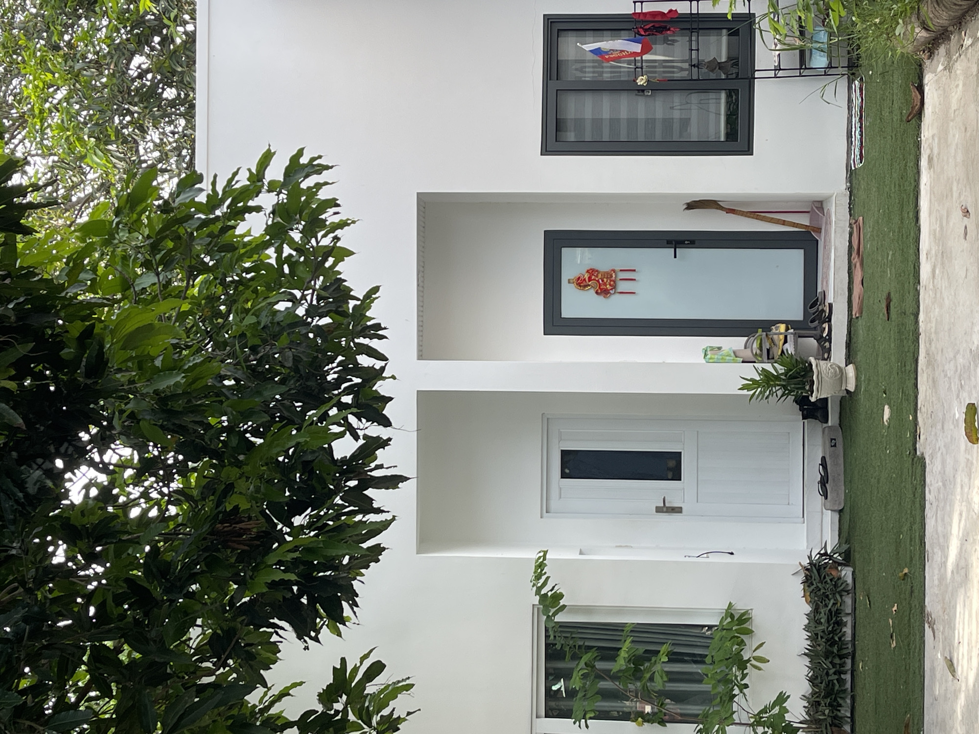 The image depicts the exterior of a modern, white building, likely a house, situated in the city of Phan Thiet Ward, Vietnam. Large green tree branches hang over the top of the structure, creating a dappled effect with sunlight.
The building features a white facade with two recessed entrances. The entrance on the left has a white door with a vertical dark panel and a silver handle. To the left of this door is a large window with a dark frame and blinds, through which a potted plant can be seen. The entrance on the right has a dark gray door with a glass panel adorned with red Chinese New Year decorations. To the right of this door is another dark-framed window with horizontal blinds.
In front of the building, a patch of artificial grass is laid out. Various objects are placed on or near the entrances and the grass. On the left side, there's a planter box filled with dark green foliage. To the right of the white door, there are a pair of black flip-flops on a grey stone. In front of the right entrance, a broom leans against the wall, and a small rug is placed on the grass. To the far right, a black metal rack holds several items, including what appears to be a Russian flag and some red fabric. A potted plant is also visible to the right of the rack.
The overall impression is one of a clean, minimalist residential exterior with some decorative elements, likely reflecting the local culture.