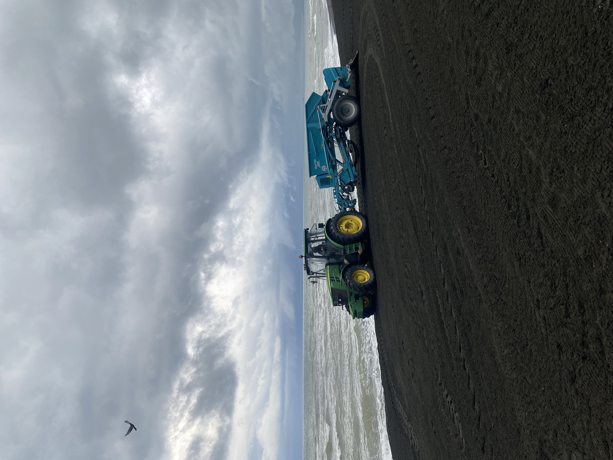 A John Deere tractor and a blue agricultural implement are parked on a black sand beach next to the choppy grey sea under a heavily overcast sky. The tractor, which appears to be a John Deere 6210R model, is green and yellow and is positioned facing right. The attached implement is a turquoise hopper with a conveyor belt and wheels, labeled "River Tech 3000". A single bird is visible in flight in the upper left quadrant of the image. The scene suggests a location in Ureki, Georgia, a town known for its magnetic black sand beaches.