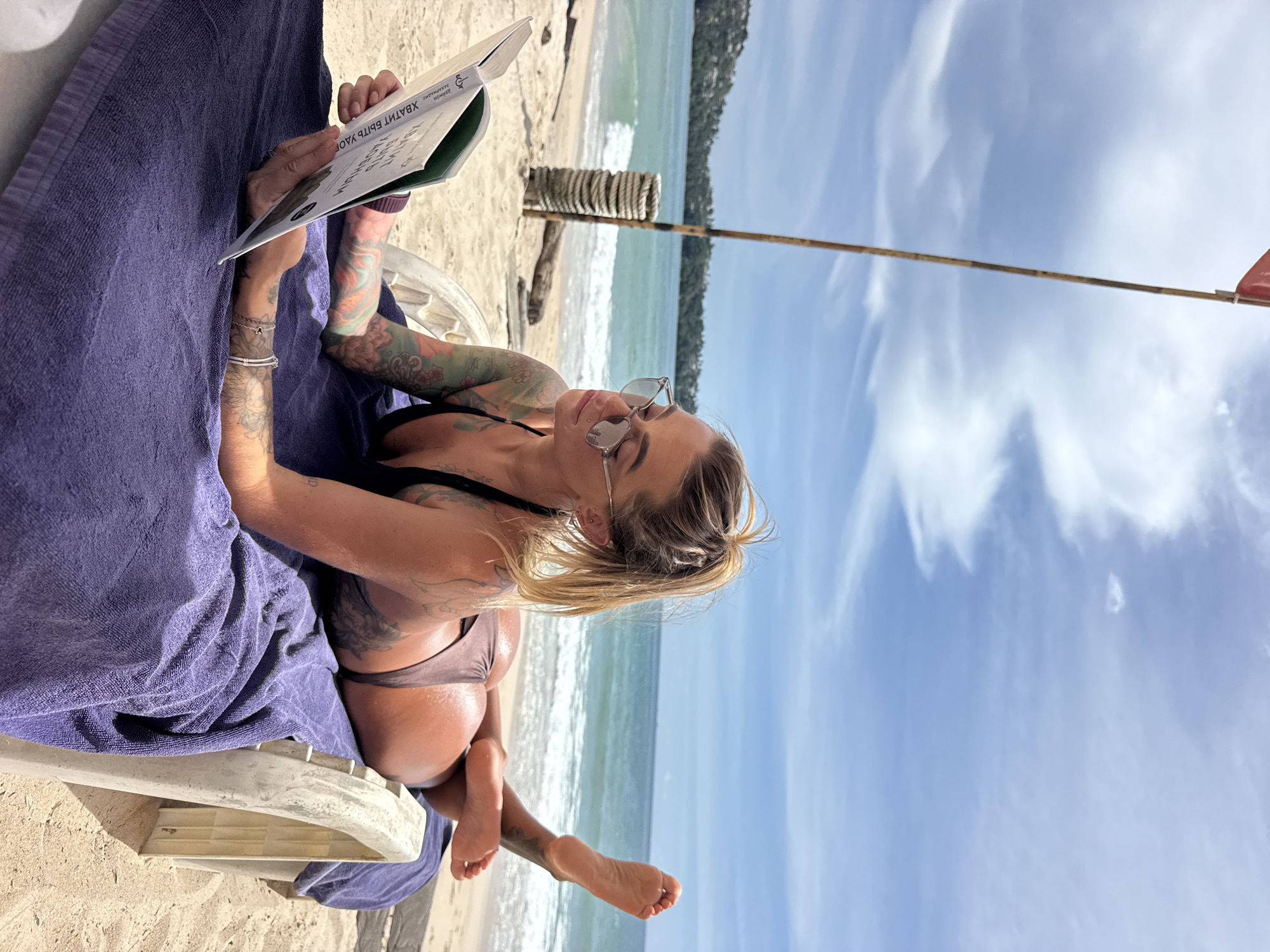 A woman is reclining on a beach chair, reading a book on a sunny day at Choeng Thale, Thailand. She has blonde hair, sunglasses, and numerous tattoos on her arms and back. She is wearing a bikini and is covered by a purple towel. The ocean is visible behind her, with waves gently lapping the shore. The sky is a bright blue with scattered white clouds. A bamboo pole with a red flag is planted in the sand nearby. The image captures a relaxed and leisurely moment, suggesting a vacation or a day spent at the beach. Text is visible on the open book in Russian, which translates to "Stop beating yourself up."