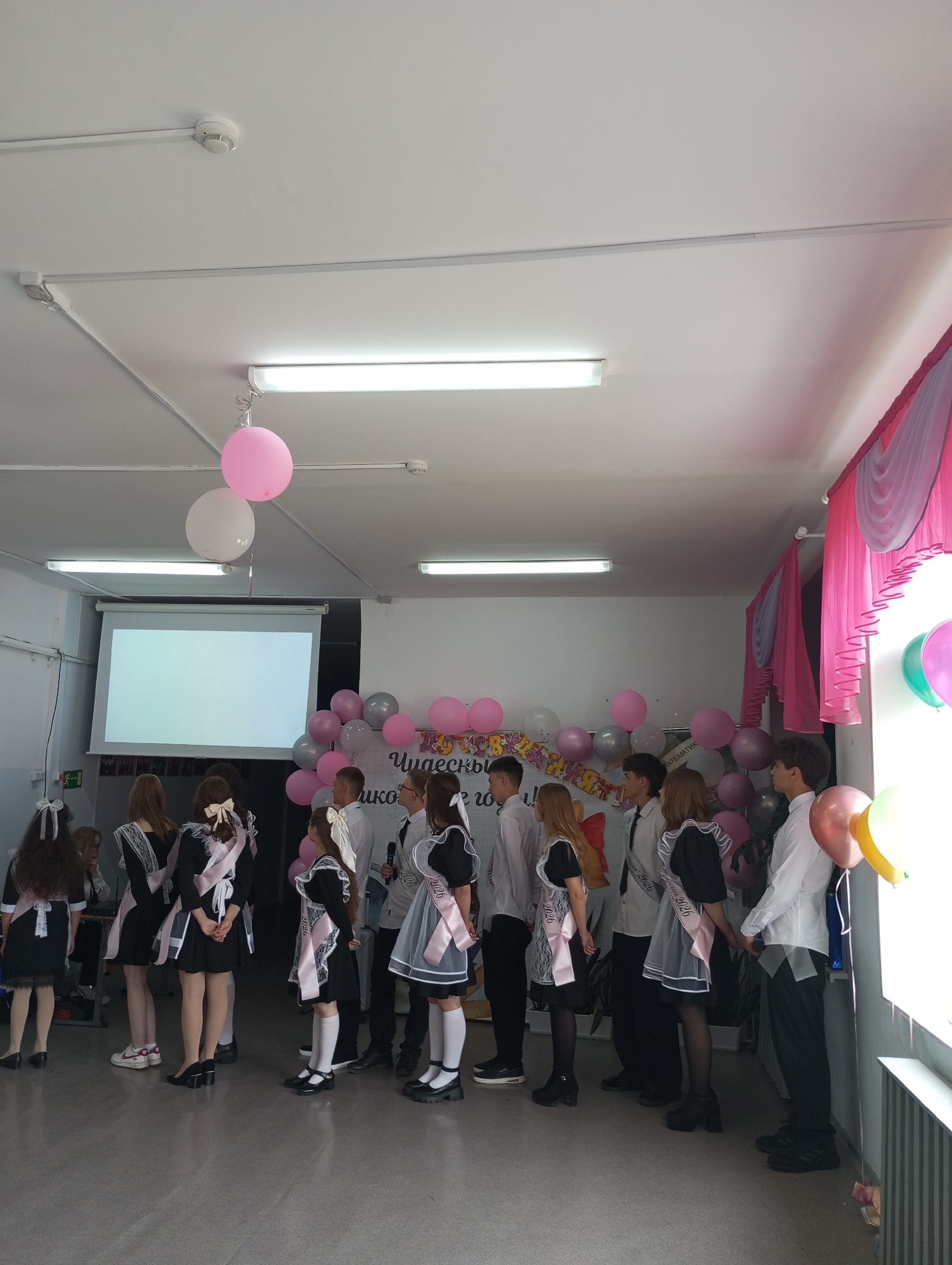 In a room in Буренка, Russia, a group of students is assembled. Most of the female students wear black dresses with white lace and ribbons, while the male students are dressed in white shirts. All students wear a sash with "2026" on it. Pink and gray balloons decorate the room. A projector screen is visible at the back, and pink curtains are draped on the right side of the room.