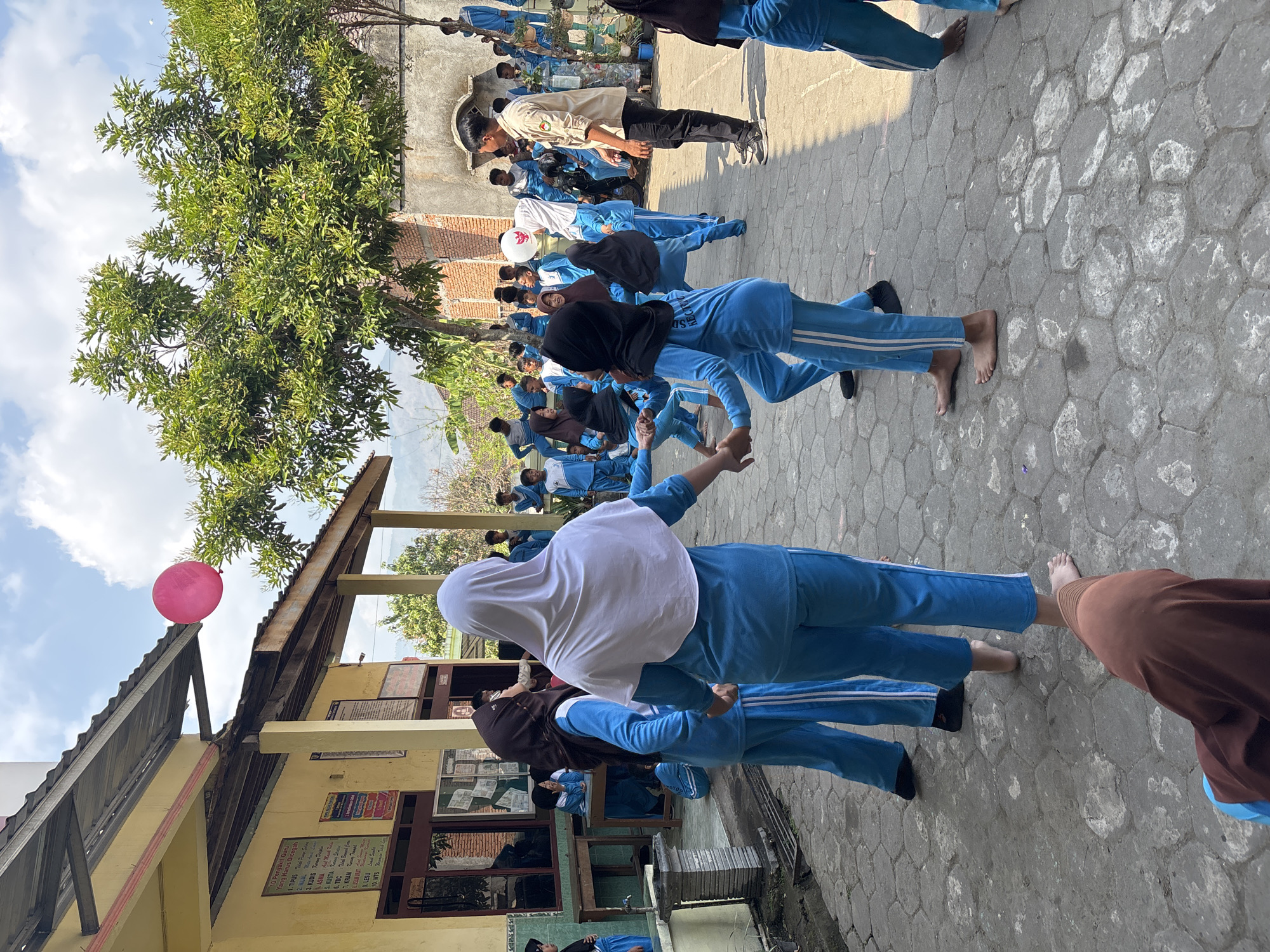 A group of children, mostly in blue uniforms, are playing outside a building with a yellow facade. A pink balloon floats in the sky above the building's roof. The children appear to be engaged in some form of group activity, with some holding hands and others observing. The ground is paved with hexagonal stones. A man in a light-colored shirt and black pants stands to the right, seemingly supervising. The environment suggests a school or community gathering. The presence of a hot air balloon and the style of the building and surrounding vegetation indicate a location in Indonesia, likely within the city of Tirtomoyo, given the provided context.