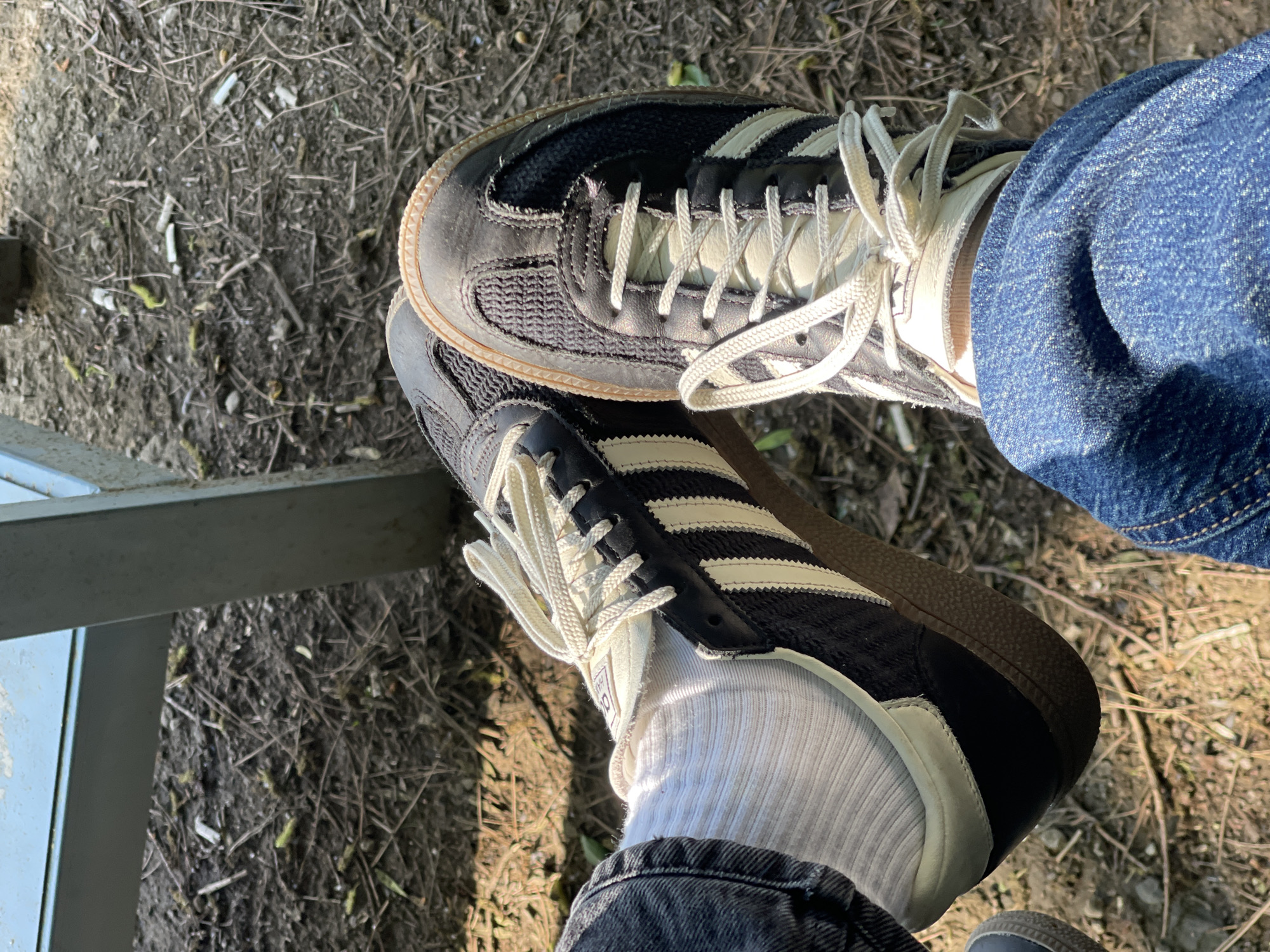 The image depicts a pair of feet wearing Adidas Samba sneakers in an outdoor setting. The upper foot is clad in blue denim jeans, while the lower foot wears dark denim jeans and white socks. The ground is covered with dirt and dry twigs. A metallic object is visible in the lower left corner of the frame. The location is not explicitly discernable from the image but is specified as Tbilisi, Georgia.