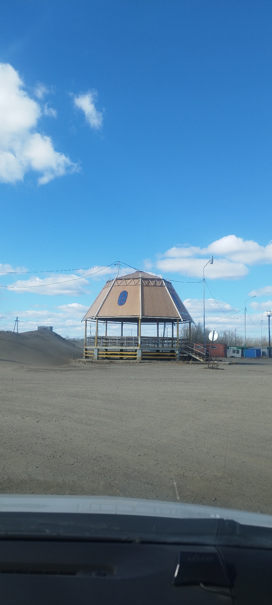 A medium shot from inside a vehicle frames a hexagonal, open-sided gazebo in Yakutsk, Russia. The gazebo has a brown, corrugated metal roof, cream-colored supporting posts, and yellow horizontal railings. It stands on a flat, barren landscape. To the left of the gazebo is a low mound of dirt, and in the background, a mixture of utility poles, street lights, shipping containers, and low buildings is visible. The sky is blue with scattered white clouds.