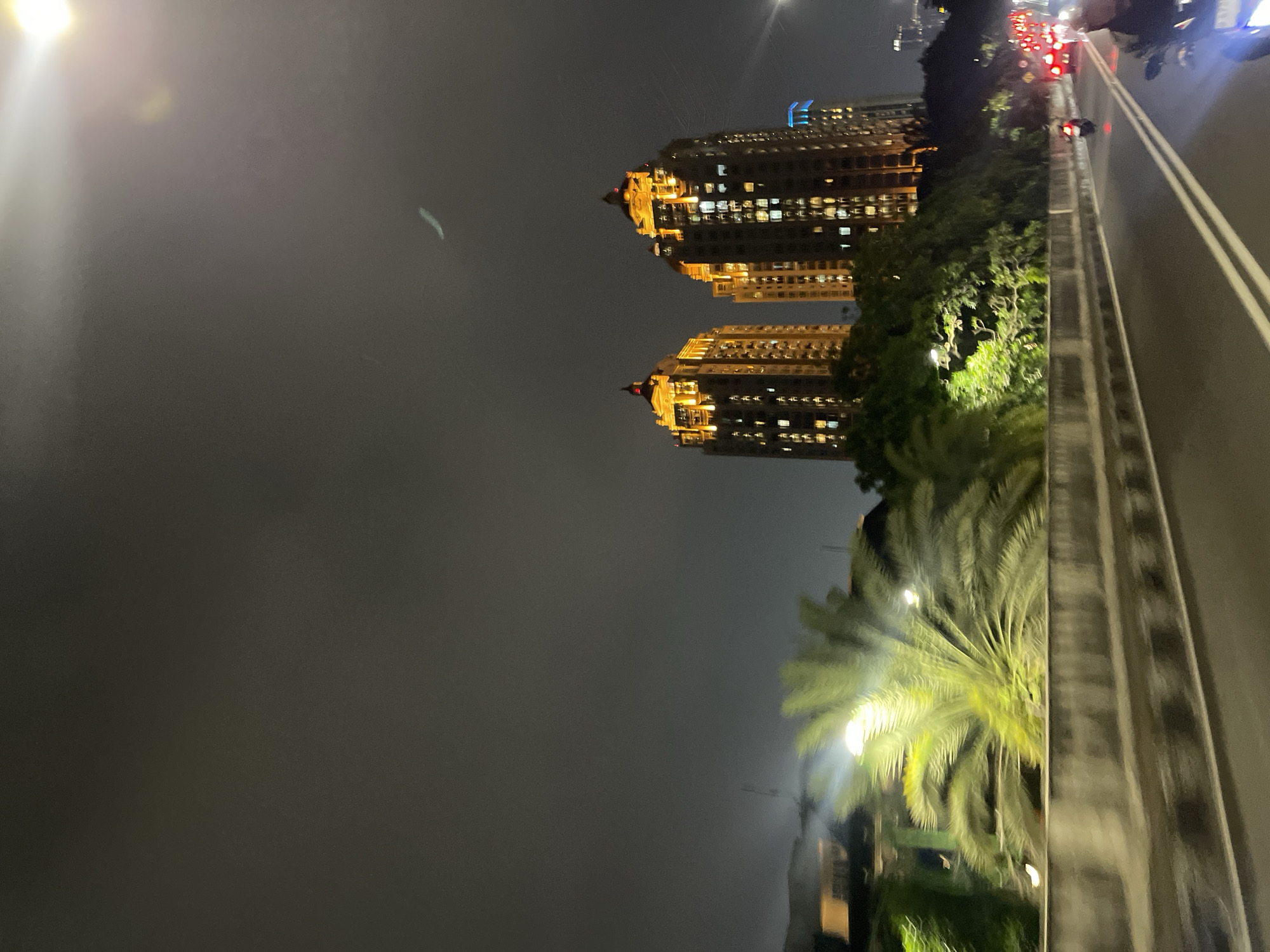 This media file captures a nighttime scene in South Jakarta, Indonesia, likely from a moving vehicle on a bridge or elevated road. The primary subjects are two tall, modern apartment buildings illuminated with warm yellow lights, creating a striking contrast against the dark, cloudy sky. The buildings appear to be twin structures, symmetrical in design and height.
In the foreground, a large palm tree with its fronds fanned out is visible, also lit by a bright light source that casts a greenish glow. The texture of the palm leaves is softened by the low light conditions and possibly motion blur. To the right of the palm tree and buildings, a road with white lane markings is visible, with several cars and motorcycles in motion, their red taillights and white headlights creating streaks of light. This suggests a busy urban environment with ongoing traffic.
The time of day is clearly night, indicated by the darkness and artificial lighting. The sky appears overcast, contributing to the overall dim and moody atmosphere. There are no visible signs of text within the image. The scene evokes a sense of urban dynamism and the vibrant nightlife of a major city, with the illuminated buildings and moving traffic suggesting activity and energy. The perspective, likely from a slightly elevated vantage point, provides a sweeping view of the cityscape.