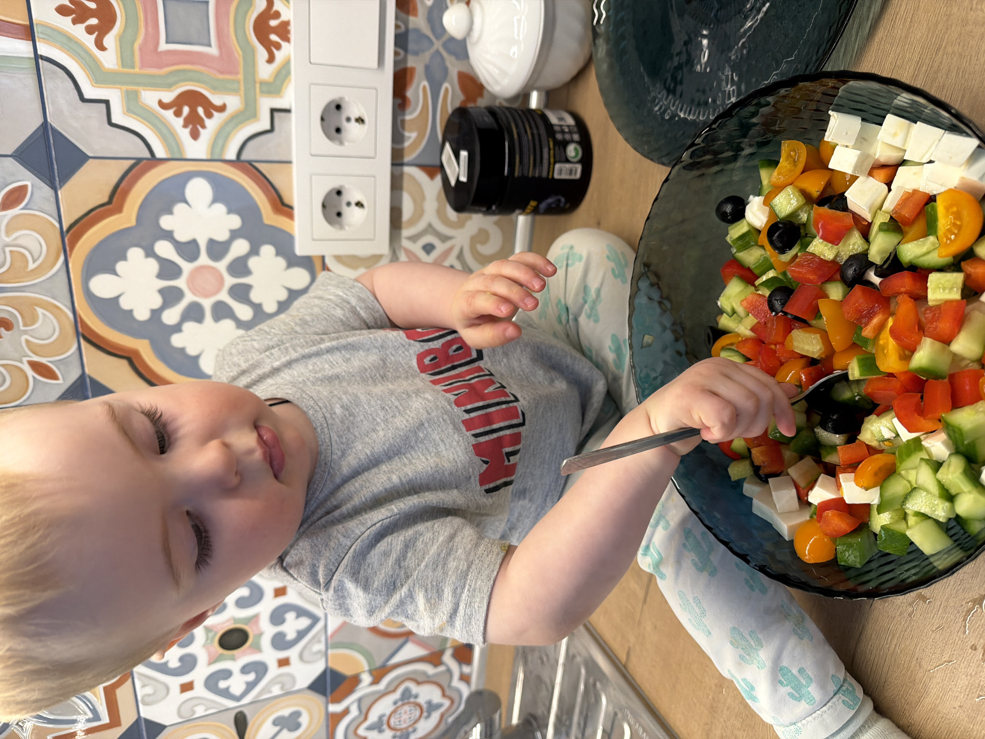A top-down, close-up shot shows a child with blond hair and fair skin, wearing a grey t-shirt with a red and black logo. The child is holding a metal spoon in their right hand, reaching into a blue-tinted glass bowl filled with a colorful salad, containing ingredients such as diced cucumbers, red and yellow bell peppers, black olives, cherry tomatoes, and cubed white cheese.
The child is sitting on what appears to be a wooden or laminate floor. To the left, there's a section of patterned tile backsplash. Behind the child, there's an electrical outlet on the wall, a white ceramic container with a lid, and a black cylindrical container with a white label.
The scene is indoors. The image provides no information to identify the location within Votkinsk, Russia.