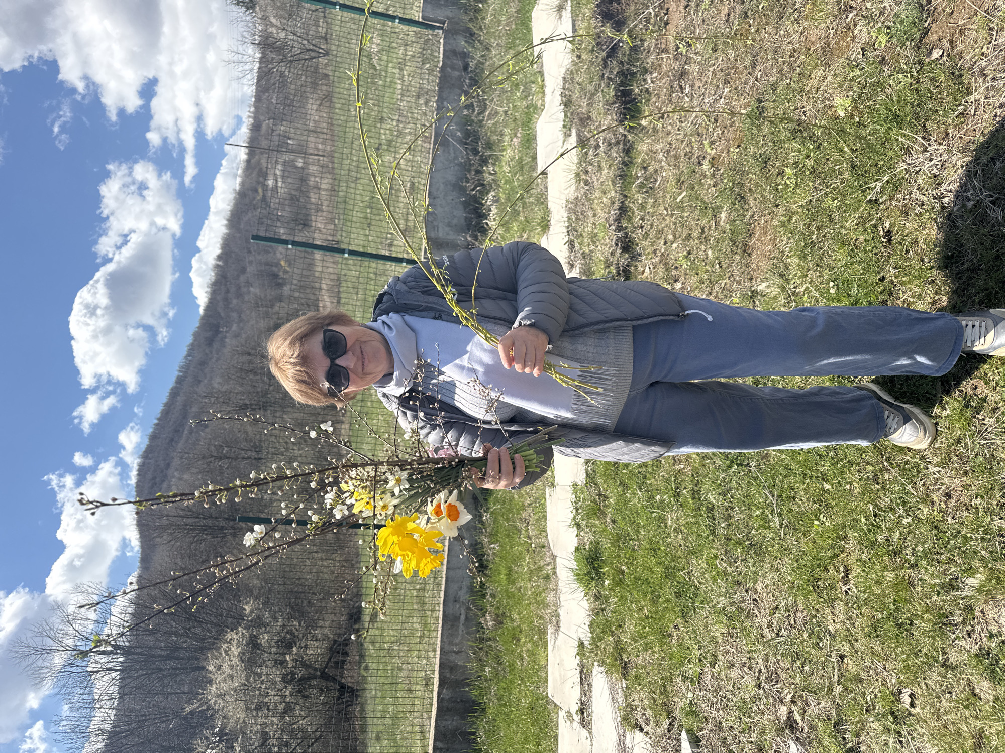 A medium outdoor shot shows a woman holding a bouquet of yellow daffodils, pussy willows, and budding branches with tiny white flowers. She stands on a grassy lawn with a mountain in the background under a partly cloudy sky. The woman wears sunglasses, a light gray jacket, a white top, and blue jeans. A green fence and concrete structures border the lawn. The location is in Unknown, Unknown.