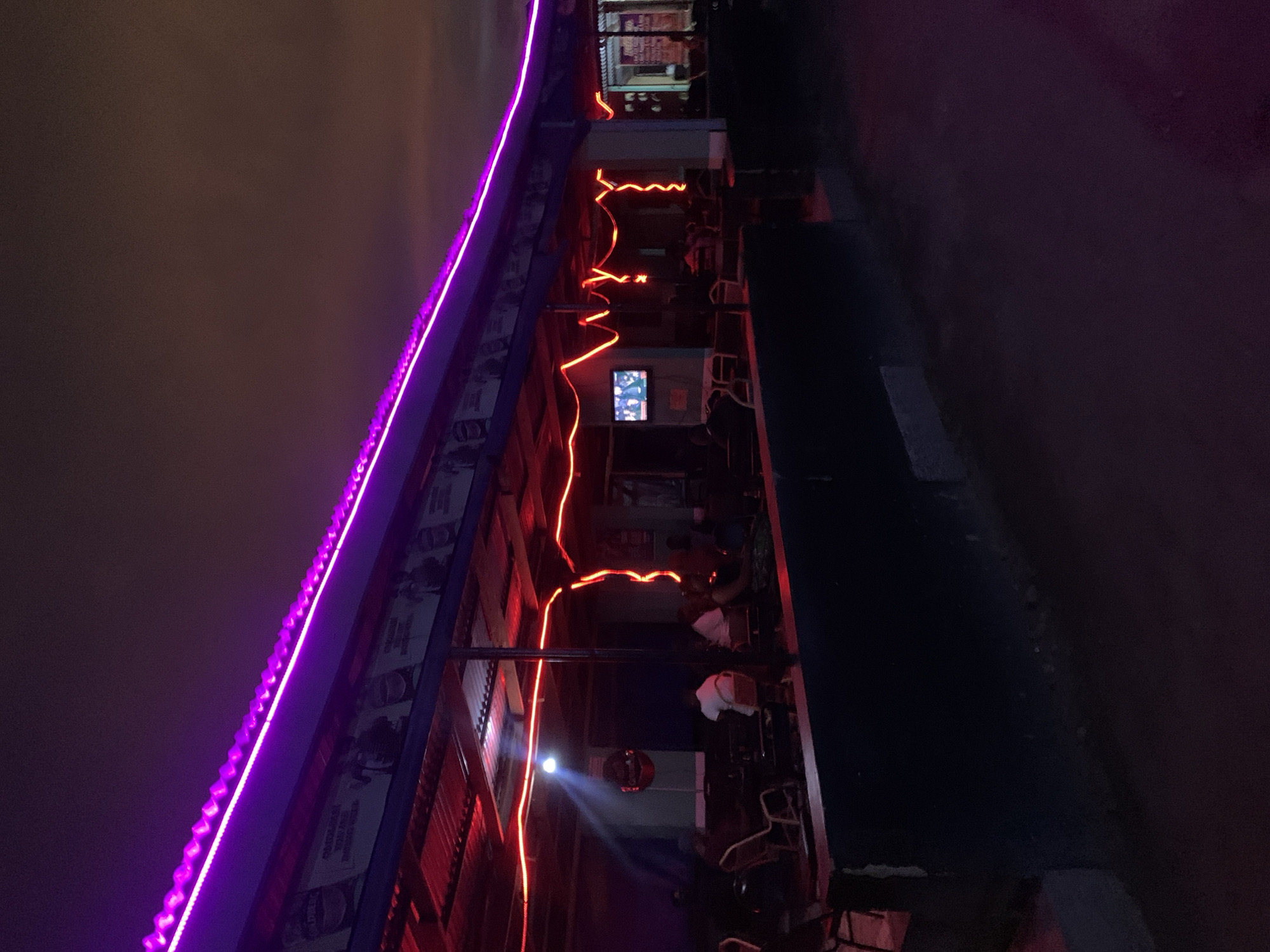 The image depicts an open-air bar or restaurant in Kabong, Nigeria, at night. The building has a long, narrow shape with a purple LED strip running along the top edge of the roof. Red neon lights zigzag across the building's façade, highlighting the seating area. Several people are seated at tables and chairs within the establishment, some appearing to watch a television mounted on the wall. A spotlight shines downward, illuminating a portion of the area. The surrounding environment is dark, with an indistinct sky overhead and a paved road adjacent to the building.