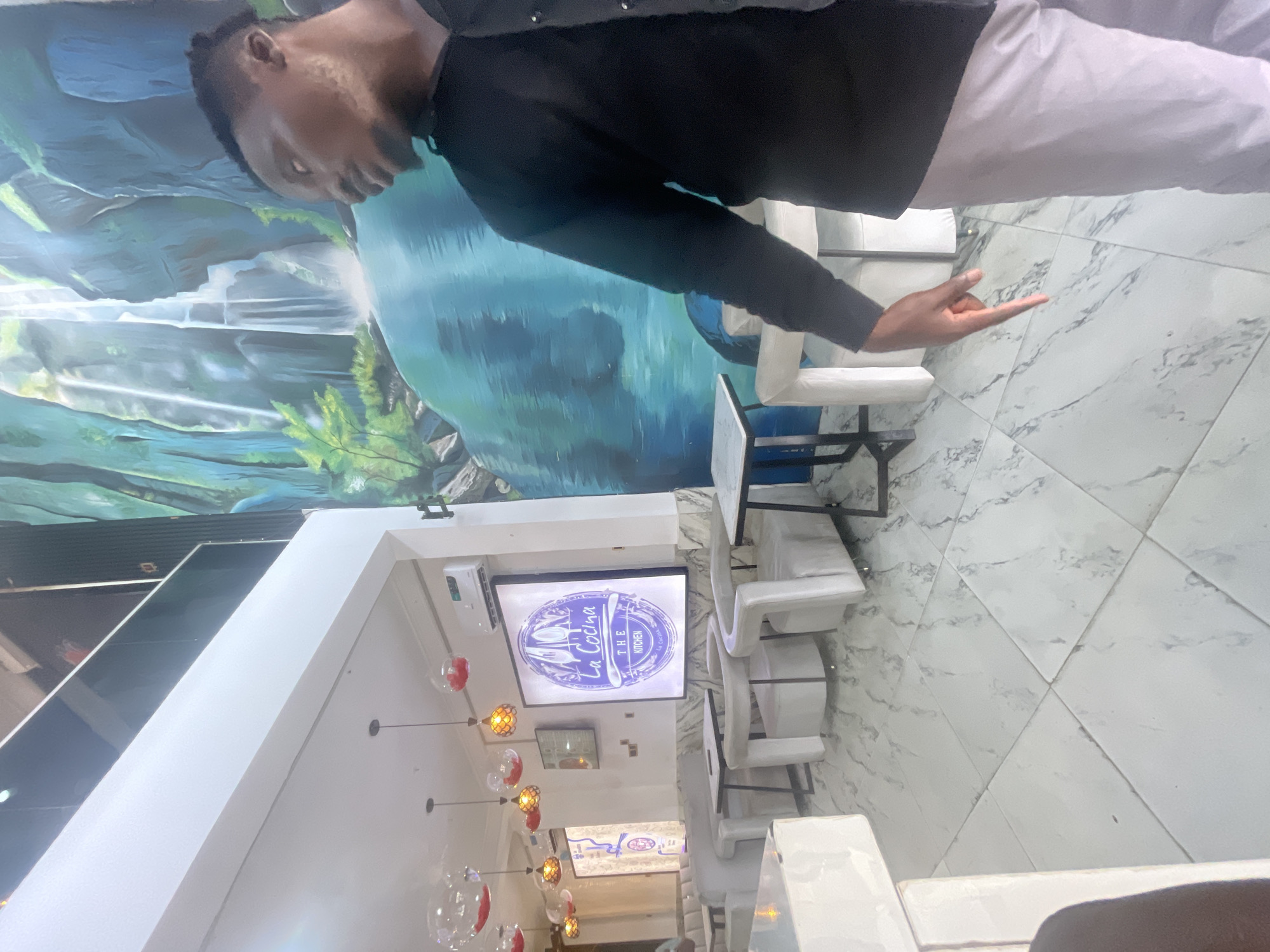 The image shows the interior of a restaurant named "La Cocina - The Kitchen," located in Ado, Nigeria. The restaurant features a modern design with white marble floors, white chairs, and pendant lighting. A large mural depicting a waterfall and nature scene adorns one of the walls. A man is standing and gesturing towards the floor. He is wearing a dark shirt and light-colored pants.