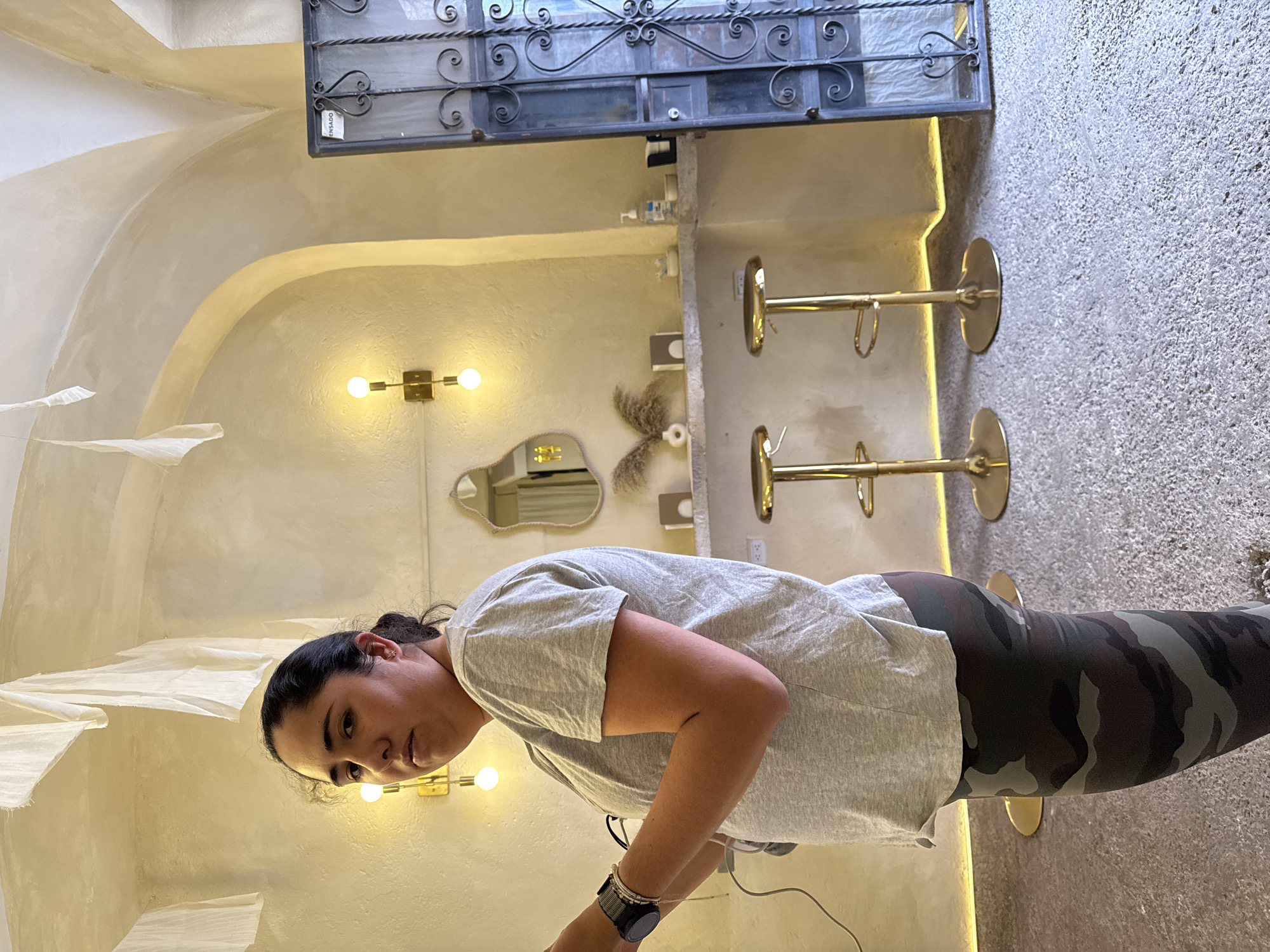 An interior shot shows a woman standing in a warmly lit, arch-filled room, likely in Puebla, Mexico. The image is oriented with the woman standing upright, though the original file is rotated. The woman, with dark hair tied back, wears a light gray t-shirt and camouflage-patterned leggings. She has a watch on her left wrist and is looking towards the camera with a neutral expression.
The room features light-colored, textured walls with soft arched recesses and a vaulted ceiling. Several wall-mounted light fixtures with exposed bulbs cast a warm glow. Decorative elements include at least two irregular-shaped mirrors and a dried floral or feathery arrangement on the wall. On the left, white, flowing fabric decorations are visible. To the right, two golden/brass-colored bar stools with round bases stand on a light, speckled concrete floor. Above, a dark metal structure with ornate scrolling, possibly a window grill or decorative panel, adds to the aesthetic. A bottle of hand sanitizer is placed on a ledge near this structure. The word "ENSADO" is partially visible on a tag attached to the metalwork. The setting appears to be a stylish cafe, bar, or hospitality space.
