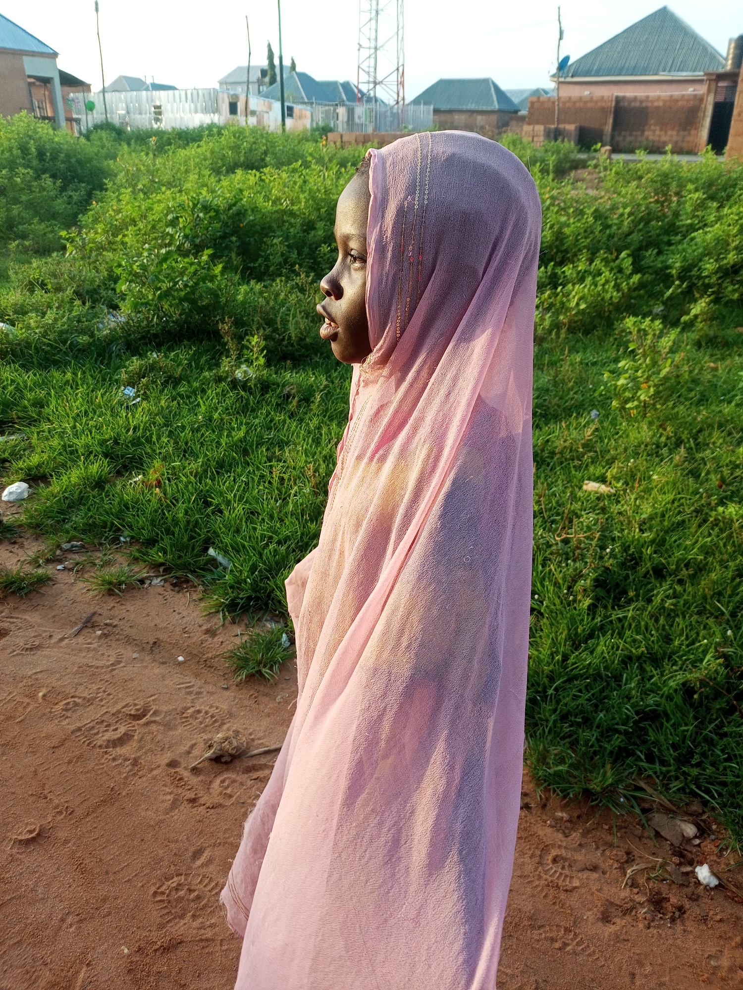 In a residential area of Tempure, Nigeria, a young girl is captured in a profile view, her head and upper body draped in a light pink, sheer hijab adorned with delicate beaded embroidery. She stands on a path of reddish-brown dirt, which is imprinted with numerous footprints. To her right, a thick expanse of vibrant green grass and foliage stretches into the background, with scattered pieces of white debris visible amongst the blades. Beyond the greenery, the scene transitions to a semi-urban landscape featuring several buildings with distinct architectural styles. On the far left, a portion of a light-colored building is visible, followed by a white wooden fence with what appears to be construction materials behind it. Further to the right, several houses with dark, pitched roofs are present, some constructed with brick and others with plastered walls. A tall telecommunications tower stands prominently in the background, adding a modern industrial element to the otherwise residential setting. The lighting suggests it is late afternoon or early evening, with the sunlight casting a warm glow on the scene.