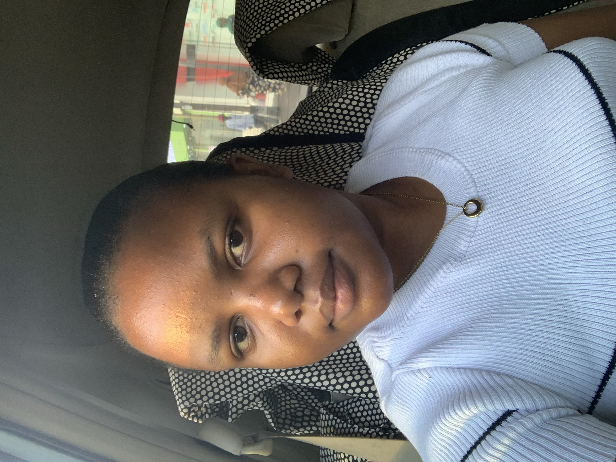 A person is in the passenger seat of a car. The person has dark hair, dark eyes, and brown skin, and is wearing a white ribbed shirt with a black trim. They are also wearing a gold necklace with a circular pendant. The seat has a black and white patterned cover. Outside the car window, people are visible on a city street in what can be presumed to be Dar es-Salaam, Tanzania.