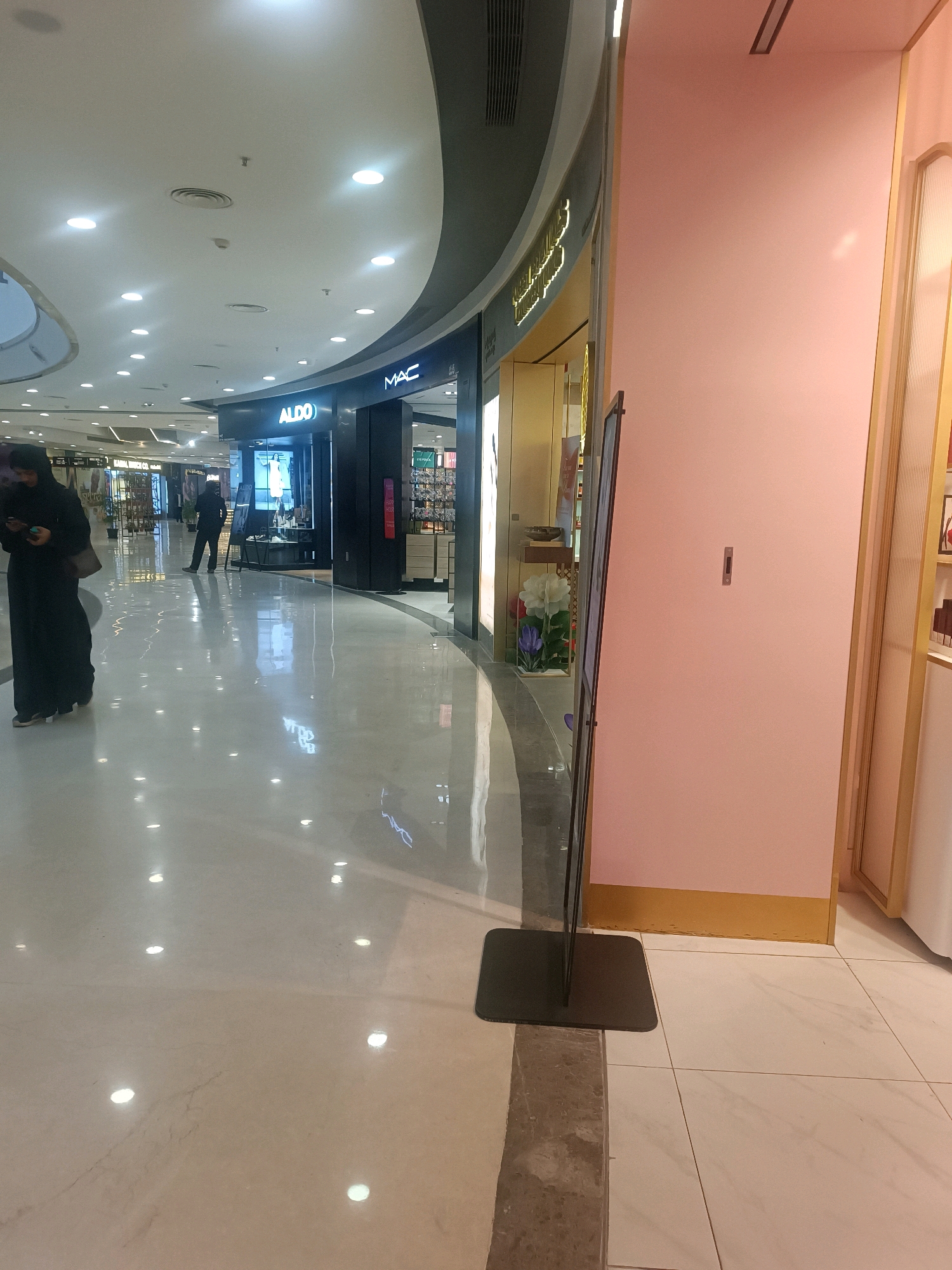 The image depicts an interior view of a modern shopping mall in Hyderabad, India. In the foreground, a woman in a black hijab is walking and looking down at her phone. The polished marble floor reflects the overhead lighting, creating a bright and airy atmosphere. Further into the mall, the storefronts of various brands, including ALDO and MAC, are visible. The architectural design features a curved ceiling with recessed lighting, typical of contemporary mall interiors. A stand with a dark-colored mat is positioned near the foreground, suggesting it might be part of a display or a temporary setup. The overall scene portrays a well-lit, clean, and active commercial environment.