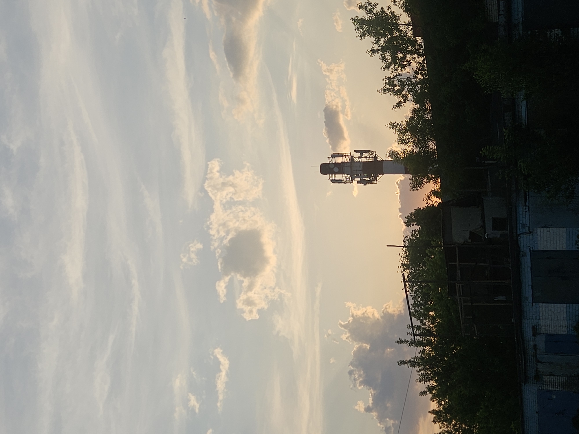 The image depicts a view of the Rodniki cityscape in Russia during sunset or sunrise, characterized by a bright, gradient-colored sky transitioning from light blue to orange. Sparse, wispy clouds are scattered across the sky. A tall industrial tower structure stands prominently against the horizon, its upper portion equipped with telecommunications apparatuses, contrasting with the adjacent, overgrown trees. At the base, rundown and semi-derelict garages are visible, suggesting an industrial or semi-industrial area. The scene evokes a sense of urban decay juxtaposed with the vast sky and technological infrastructure. There are no people visible in the frame.