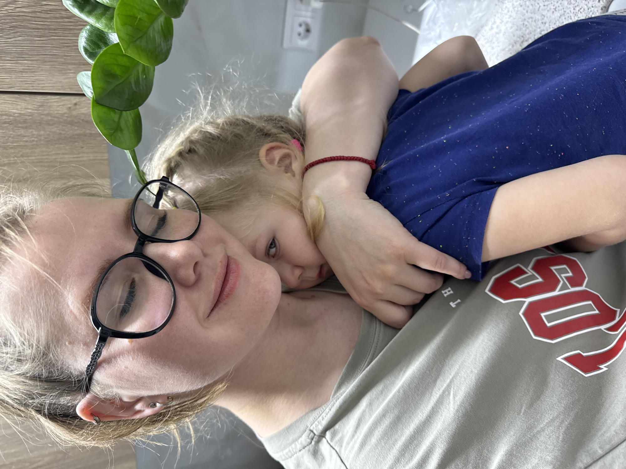 This image, set in "Ферма, Russia", captures a close-up, tilted view of an adult and a young child in a tender embrace indoors. On the left, an adult, likely female, with blonde hair and black-framed glasses, rests her head, her eyes appearing gently closed or cast downwards. She wears a light olive-green t-shirt with partial white letters "TH" and red and white graphic elements resembling "SO" or "OS" visible on the chest area. Her ear shows multiple piercings.
Nestled intimately against the adult's shoulder and neck is a blonde-haired child, possibly female, with pigtails secured by a pink hair tie. The child, wearing a dark blue t-shirt with colorful speckles, looks calmly towards the camera. Her right arm is wrapped around the adult's neck, and a red string bracelet is visible on her wrist.
The background suggests a casual home environment, featuring a light wooden surface on the left and a pale wall with a white electrical outlet. A vibrant green houseplant is partially visible in the upper left corner. The scene conveys a warm and affectionate moment of comfort or rest between the two subjects.