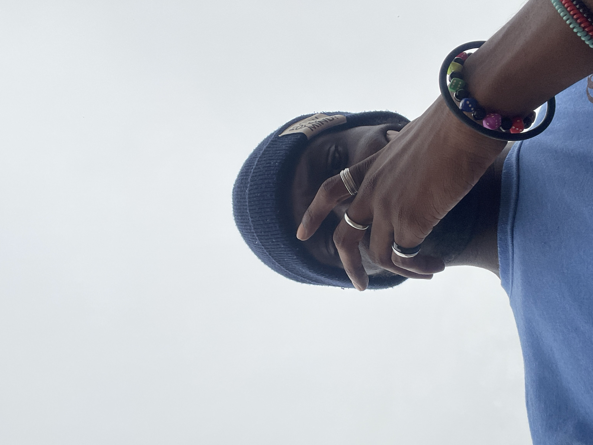 The image shows a low-angle, close-up shot of a person from the chest up, wearing a blue t-shirt and a dark blue beanie. The person's right hand is raised, partially covering their face. Several rings are visible on their fingers, including two on the ring finger and one on the middle finger of the right hand. The person is also wearing a colorful beaded bracelet and a black rubber bracelet on their right wrist. The background is a solid, overcast sky, suggesting an outdoor setting. The image's composition and the absence of specific landmarks make it impossible to determine the exact location within Abuja, Nigeria.